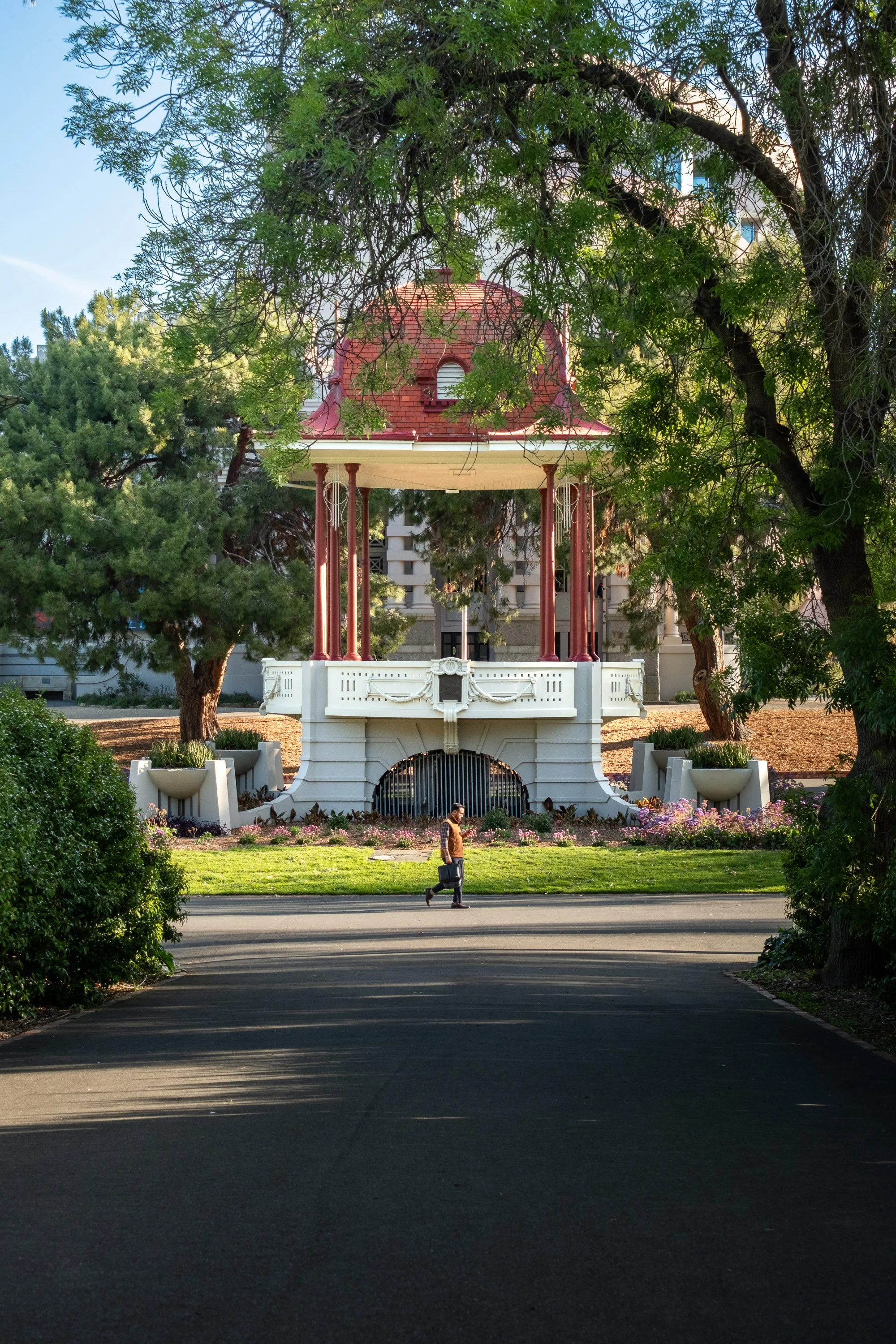 A man with a suitcase walking on a paved path towards a decorative gazebo with a red roof and columns, surrounded by trees and flowers.