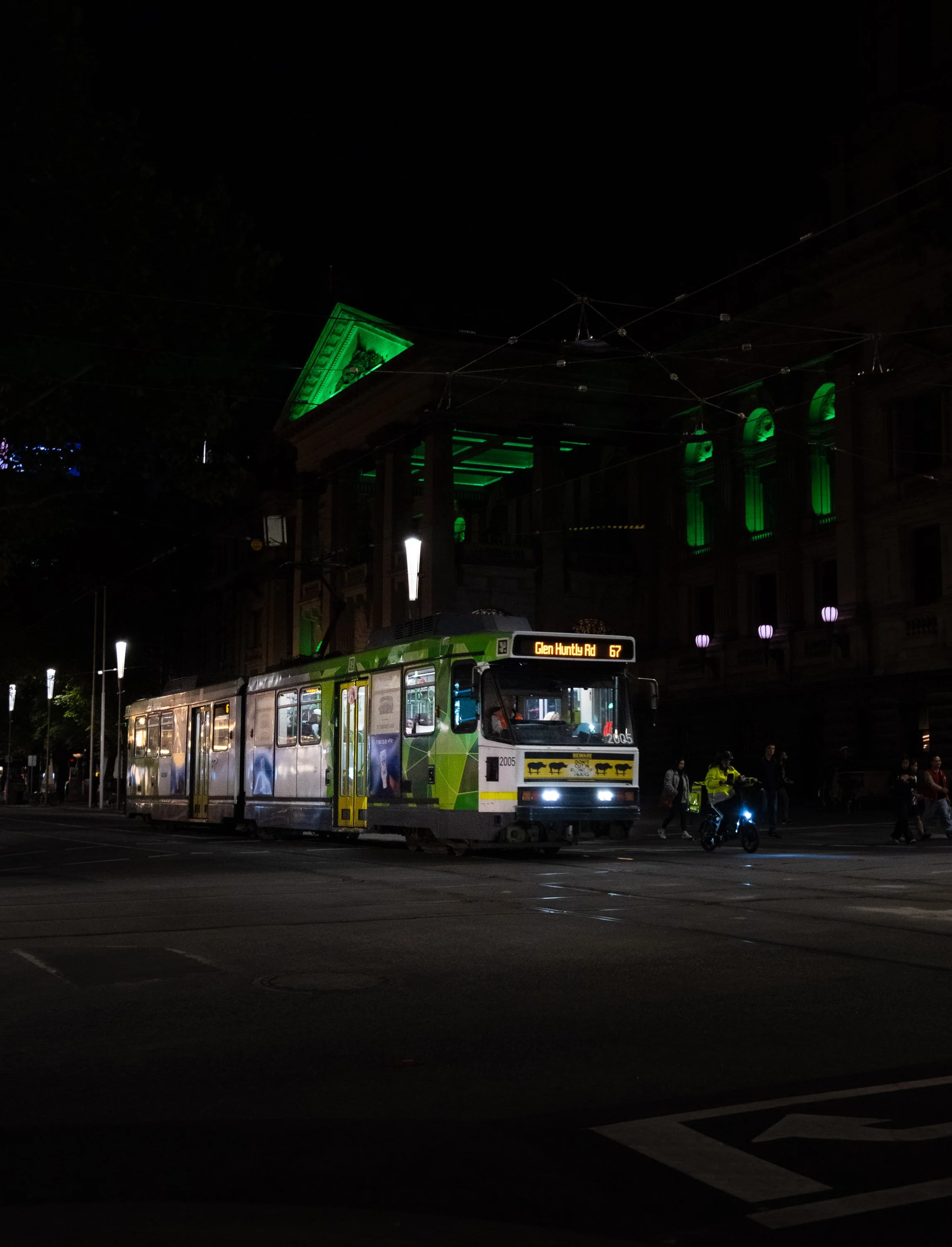 A tram at night in an urban area with an electronic sign showing 'Glen Huntly Rd 67'. The tram is green and white, with advertisements on the side, and is moving across a street with some people and a motorcyclist nearby. In the background, a buildin