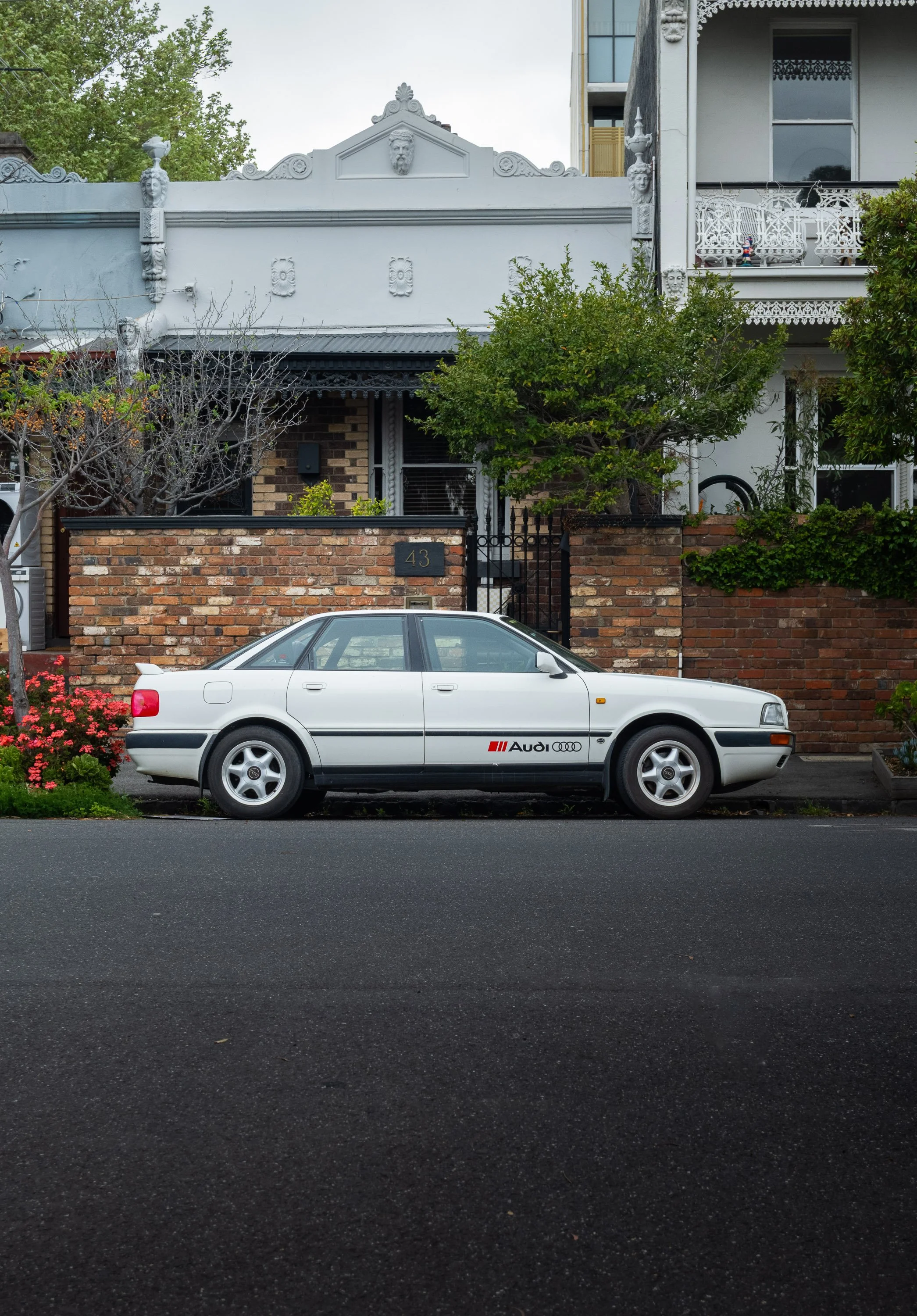 A white vintage Audi car parked on the street in front of a residential area with brick and ornate Victorian style houses and greenery.