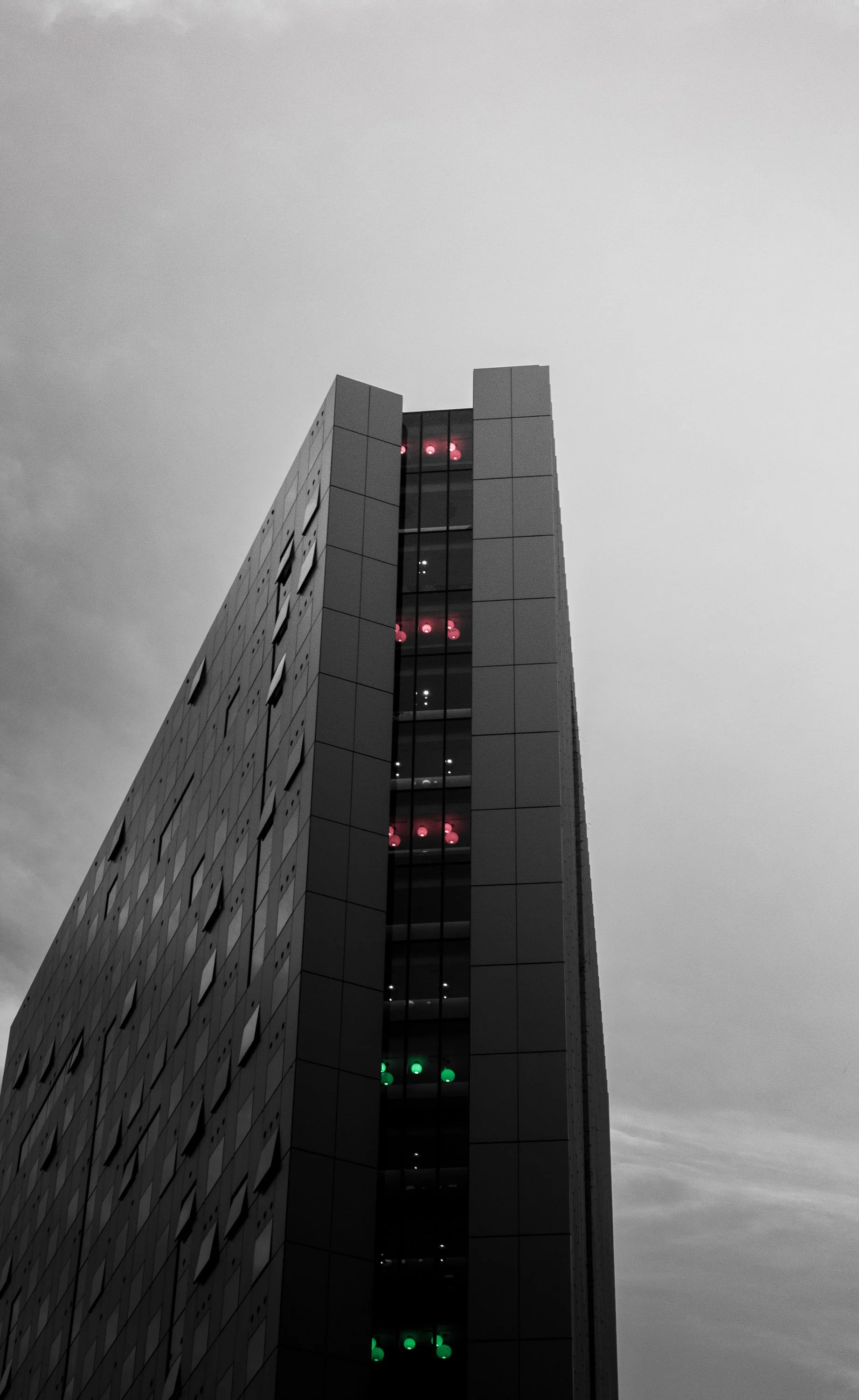 Tall modern building with illuminated green and red lights on the corners of its glass elevator shaft against a cloudy sky.