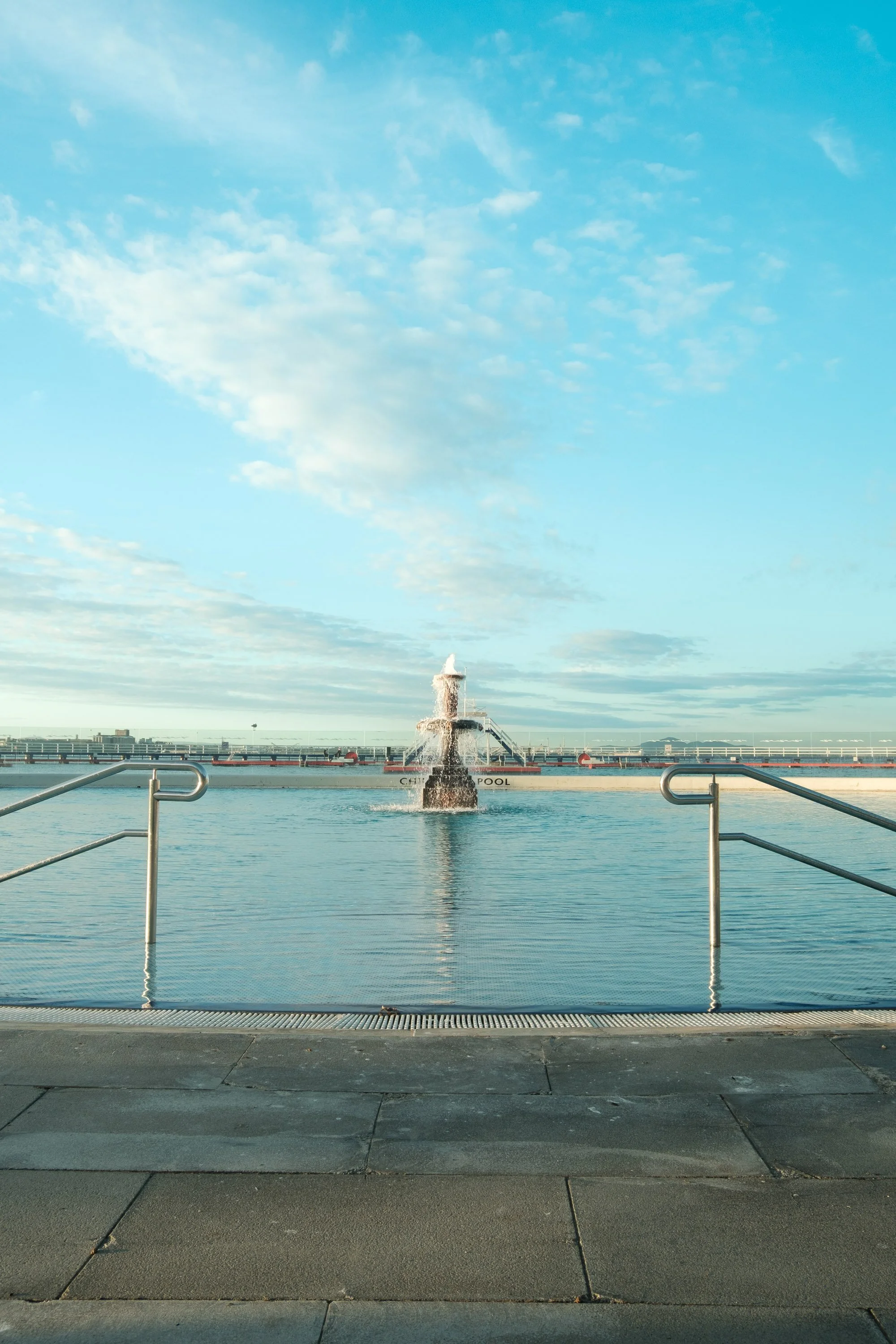 Fountain with a water jet in a swimming pool, metal railings on either side, and cloudy sky in the background.