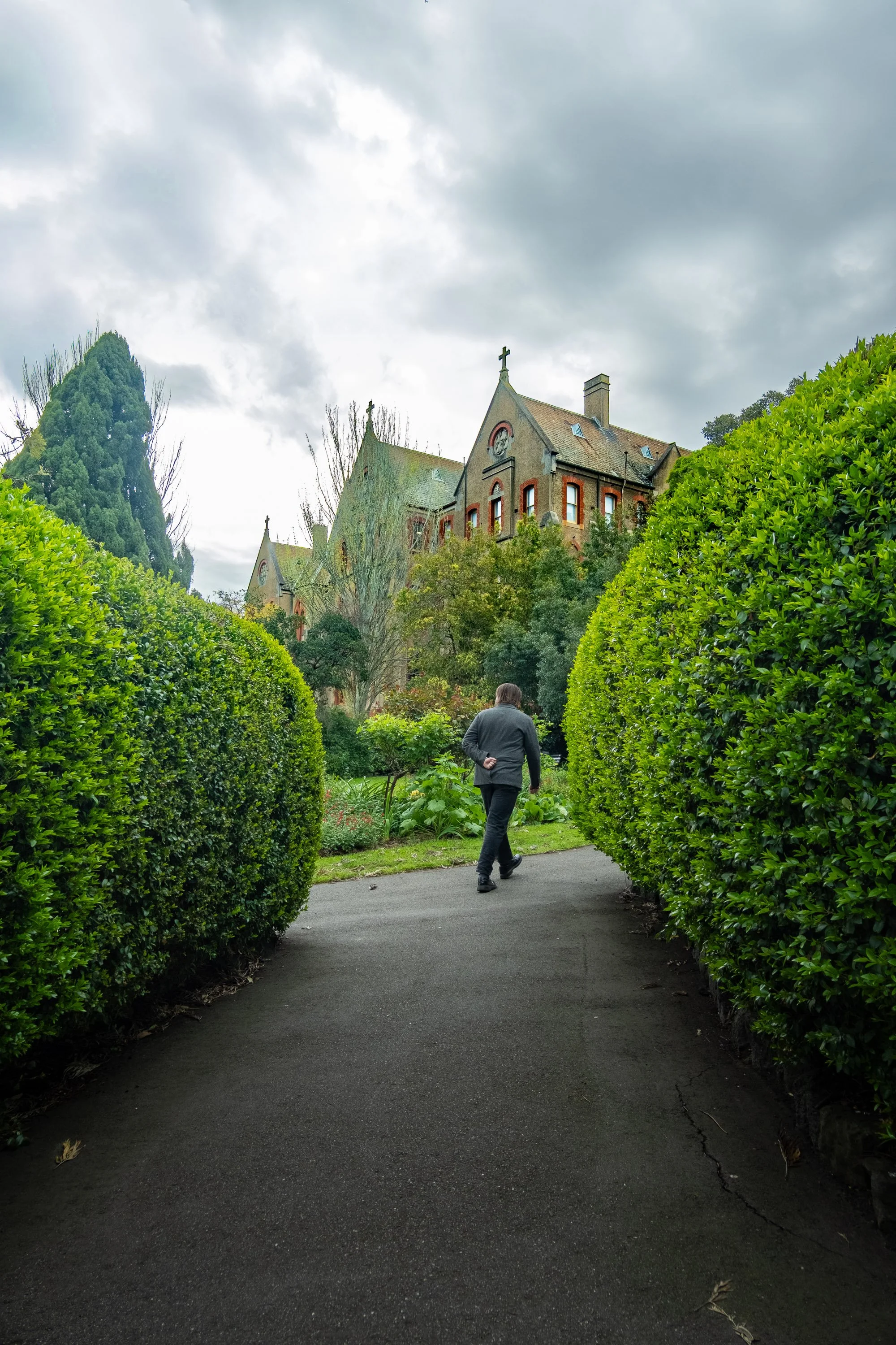 A person walking on a paved path through a lush garden with neatly trimmed bushes, leading towards a large historic brick building with Gothic architectural features, under a cloudy sky.