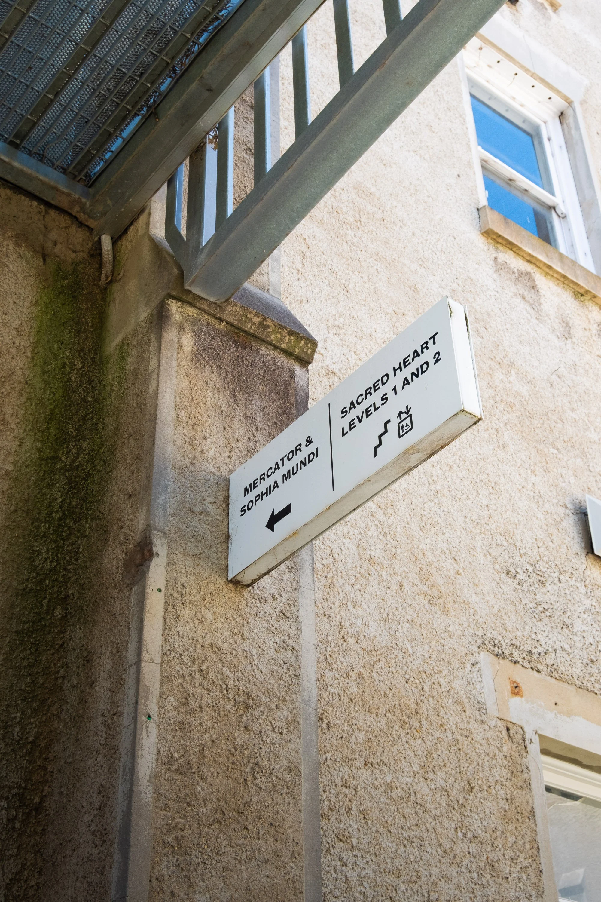 Street sign indicating directions to 'Mercator & Sophia Mundi' to the left and 'Sacred Heart Levels 1 and 2' upstairs, on a beige stucco wall.