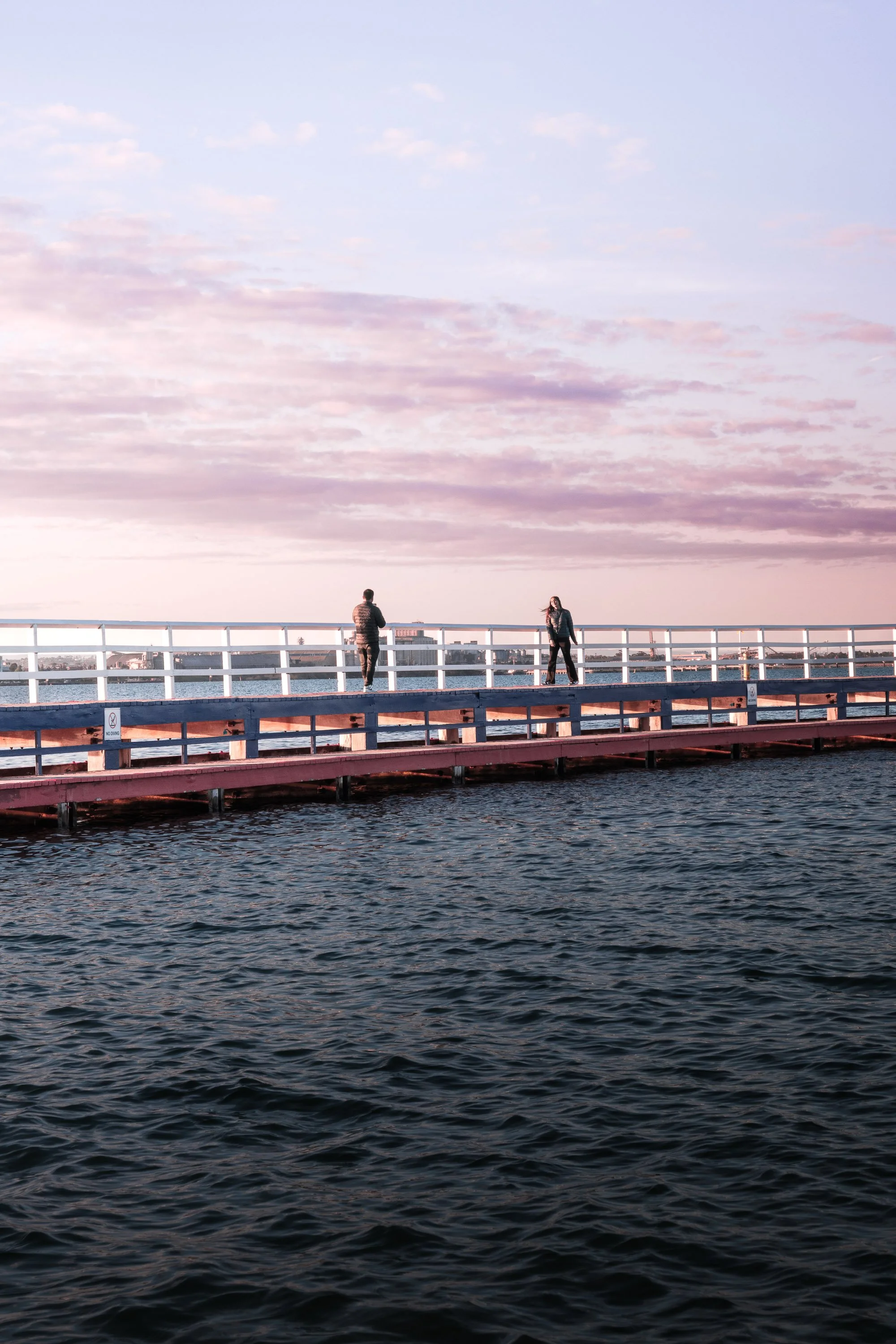 Two people standing on a pink and white dock over dark water, with a sky full of soft pink and purple clouds in the background.