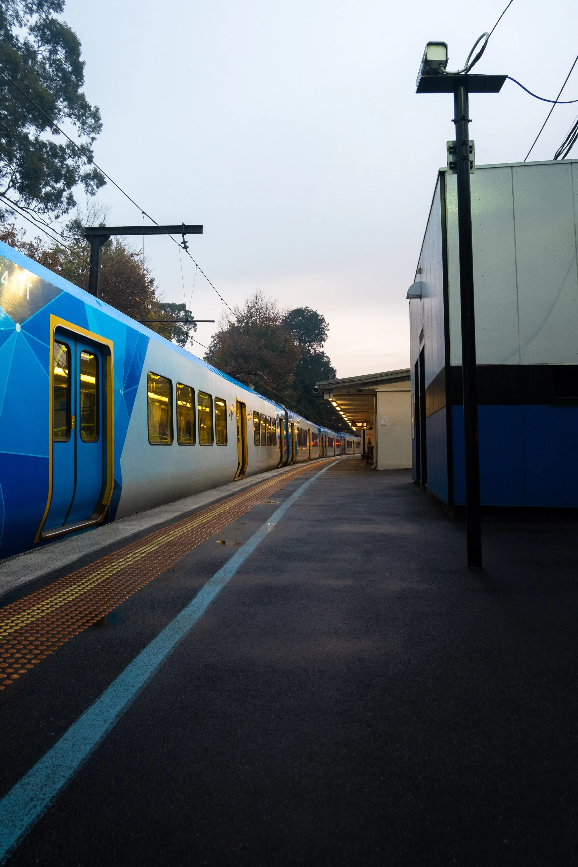 A train at a station platform during dusk with trees in the background and a pole with electrical wires overhead.