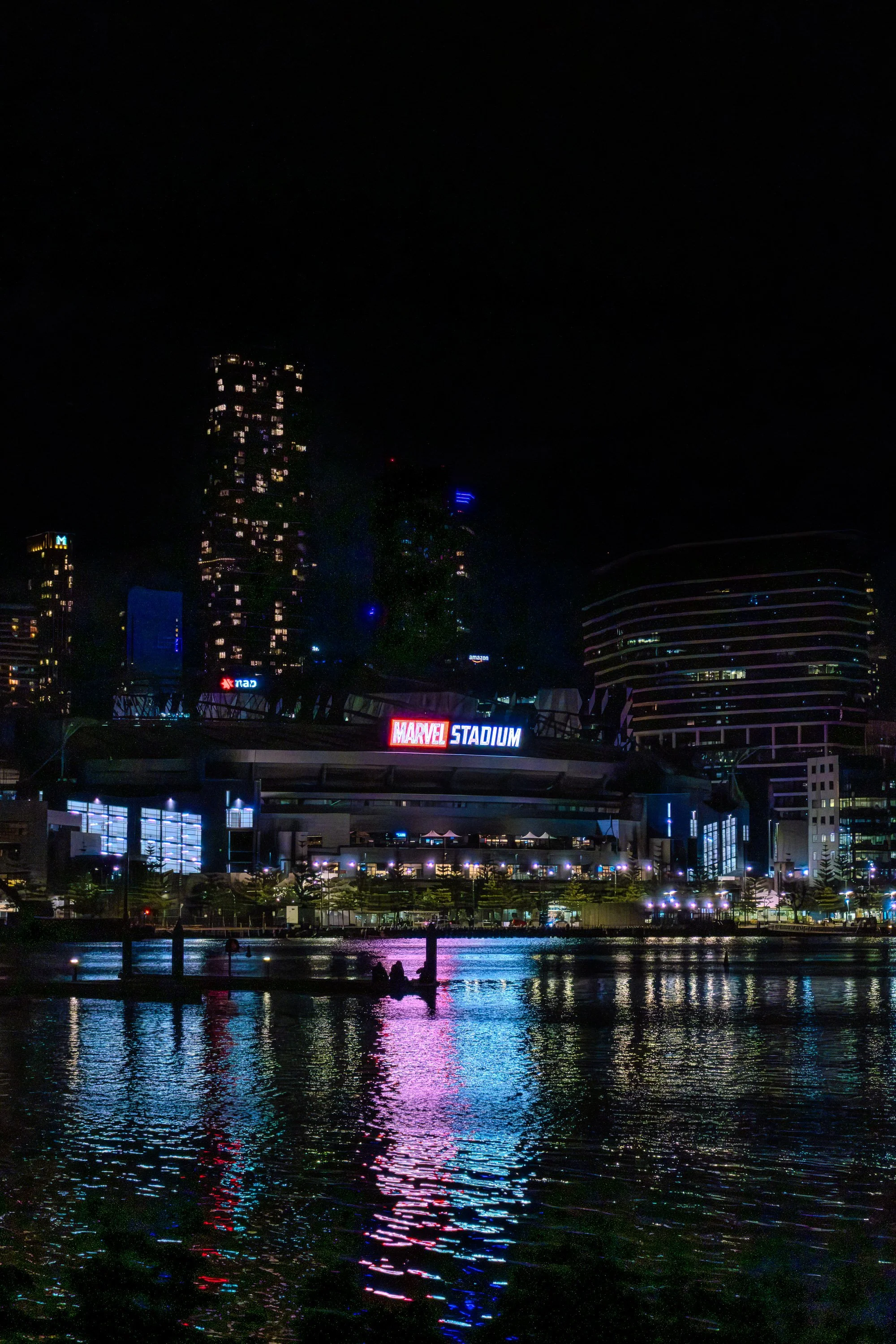 Nighttime view of Marvel Stadium with city skyline in the background, lights reflecting on the water in front.