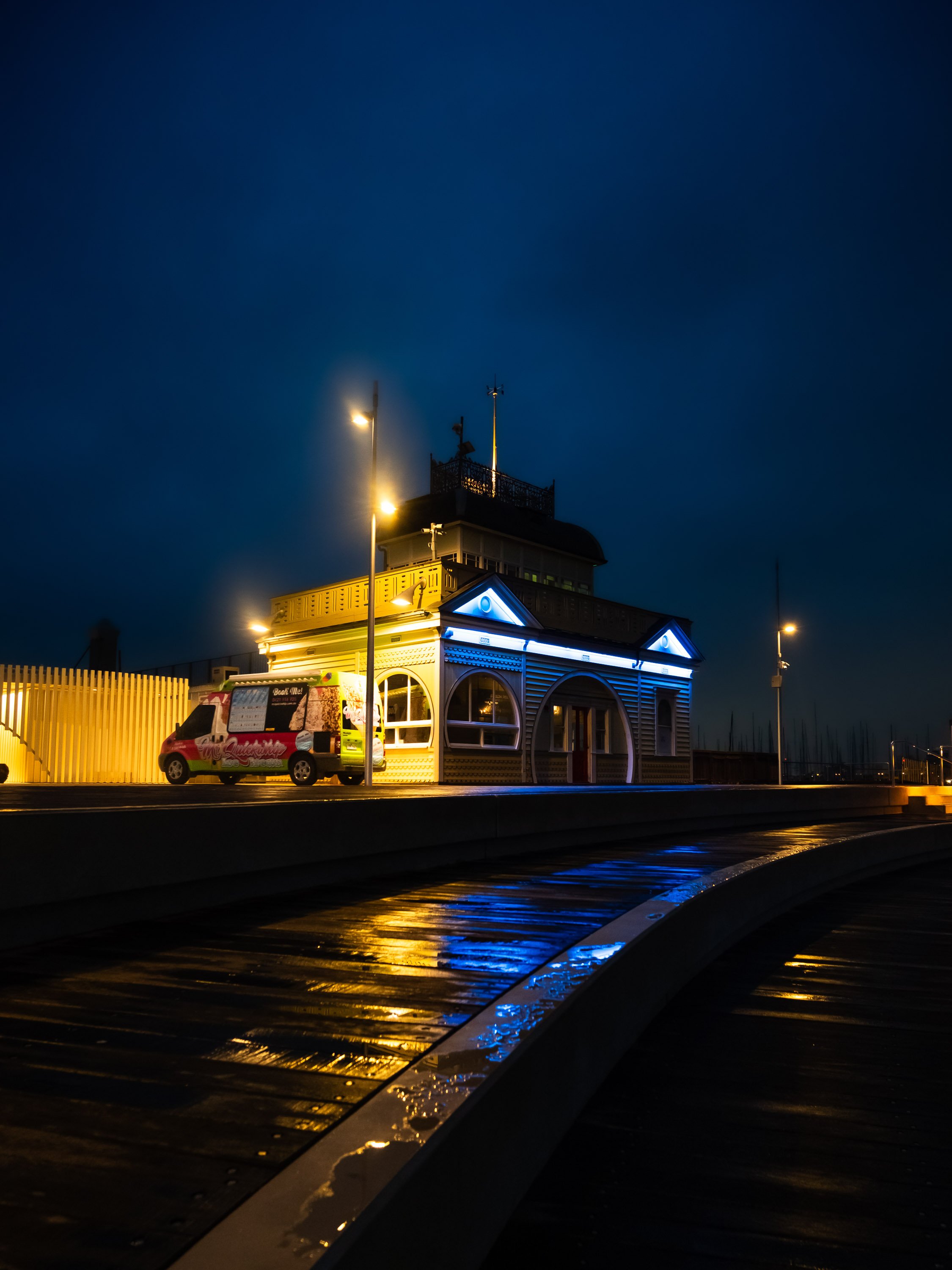 A brightly lit building with arched windows and decorative trim at night, with a colorful van parked outside and streetlights illuminating the scene. The wet ground reflects the lights.