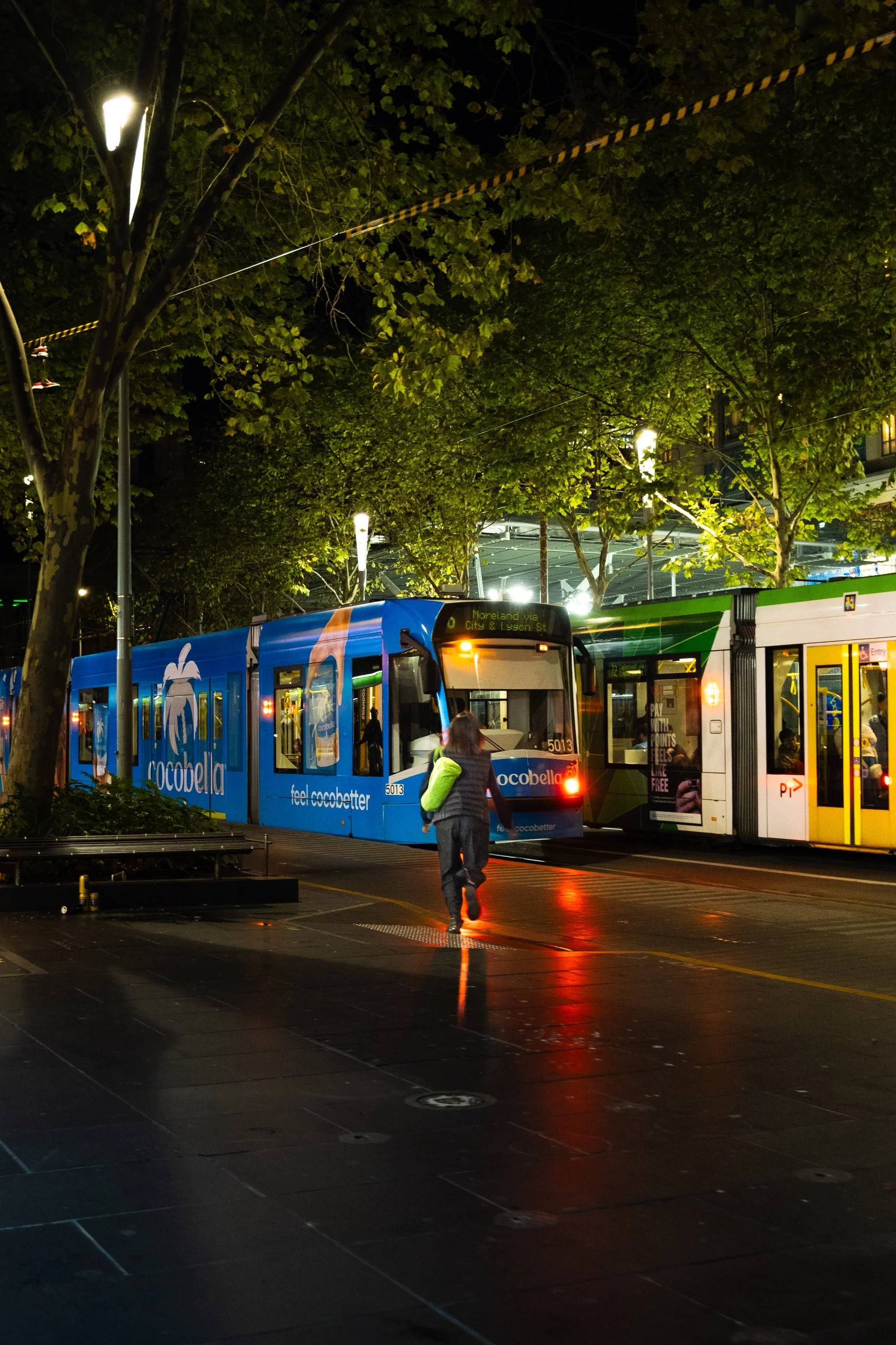 Night scene of a city street with two light rail trams stopped at a station, one blue and one white with green, under a canopy of trees illuminated by streetlights. A woman in a dark jacket and jeans walks towards the blue tram, which has an advertis