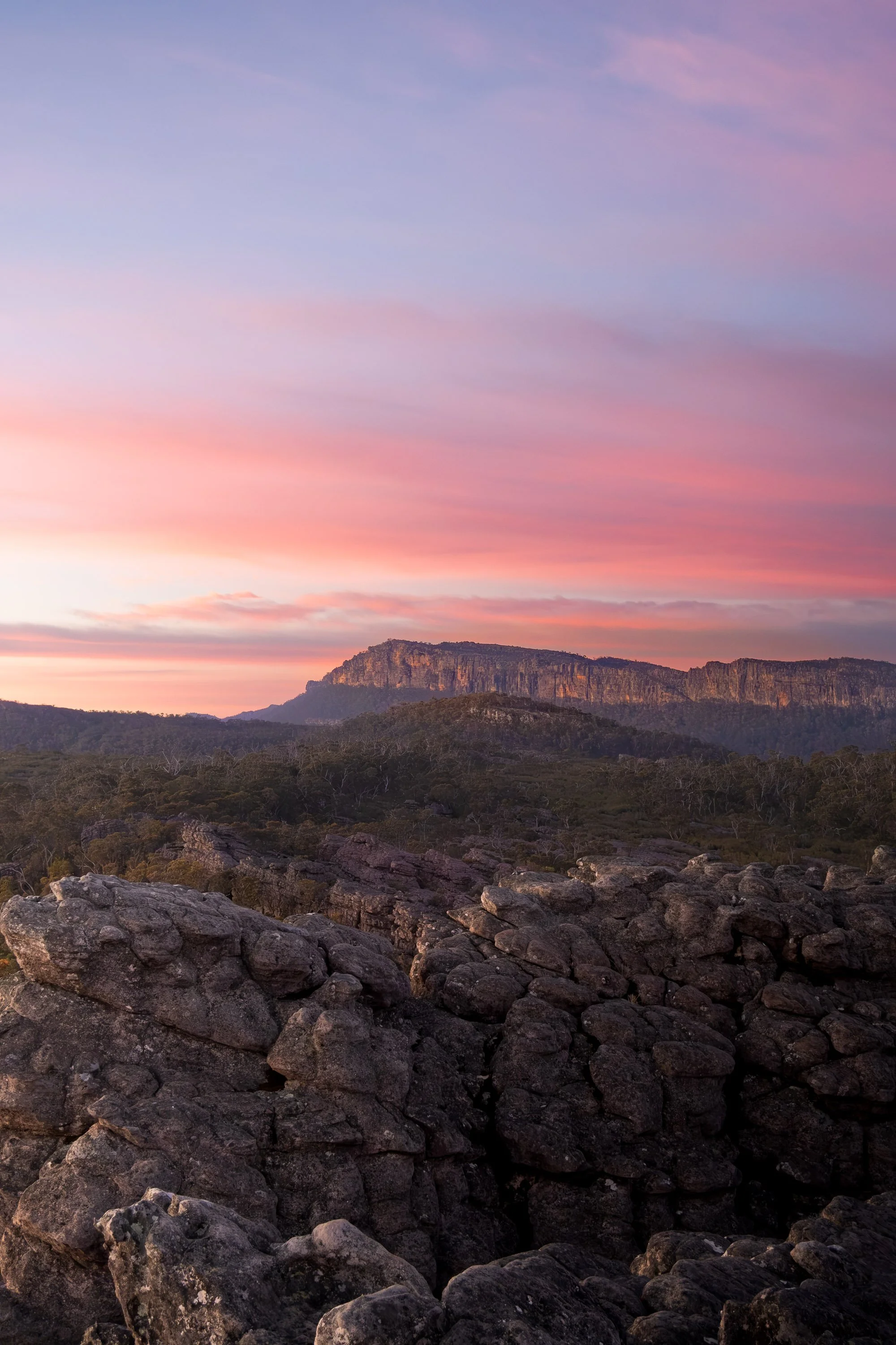 A scenic landscape at sunset showing rocky terrain in the foreground, dense forest in the middle ground, and a mountain range in the background under a colorful sky with pink and purple hues.