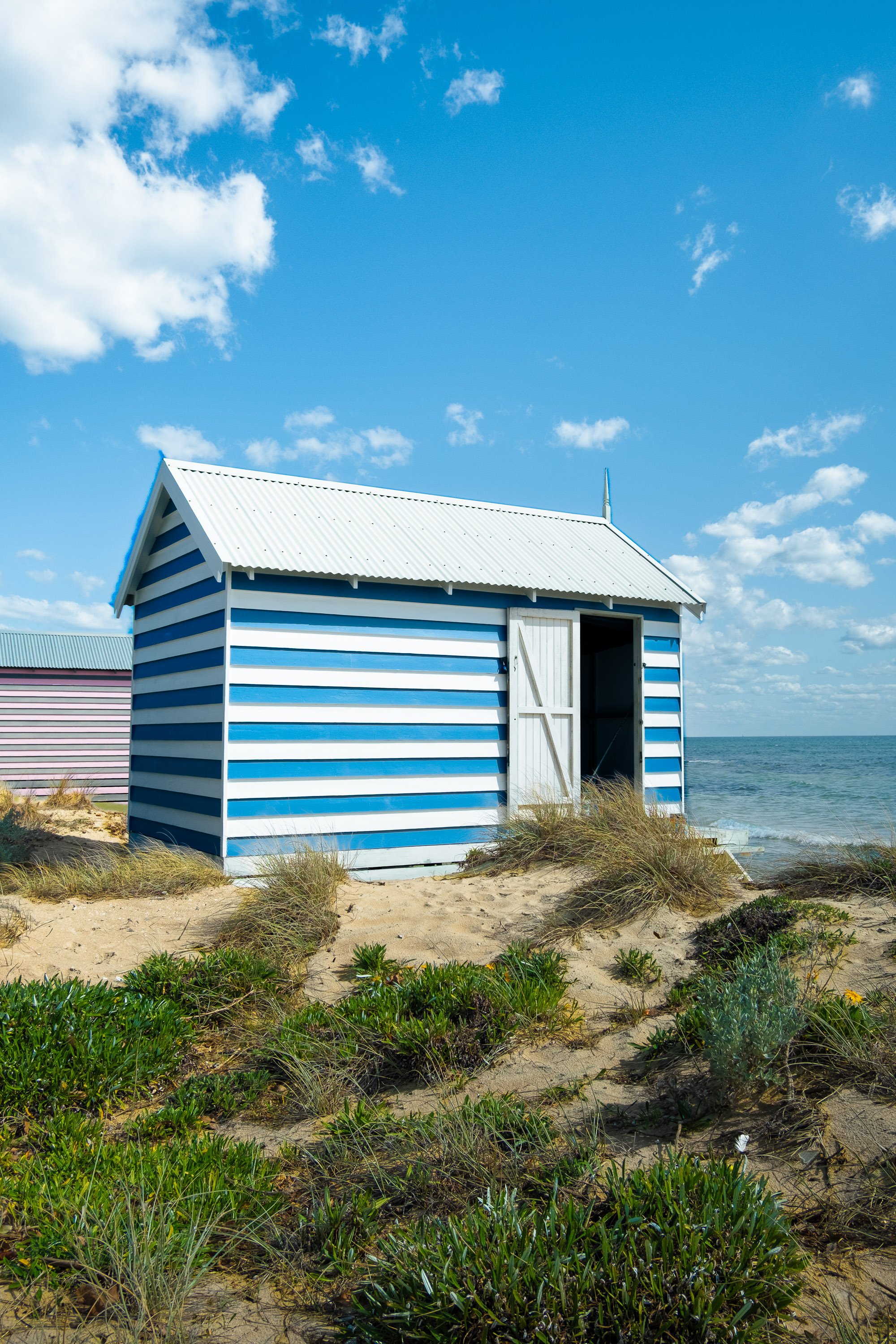 A blue and white striped beach hut on sandy dunes near the ocean under a partly cloudy sky.