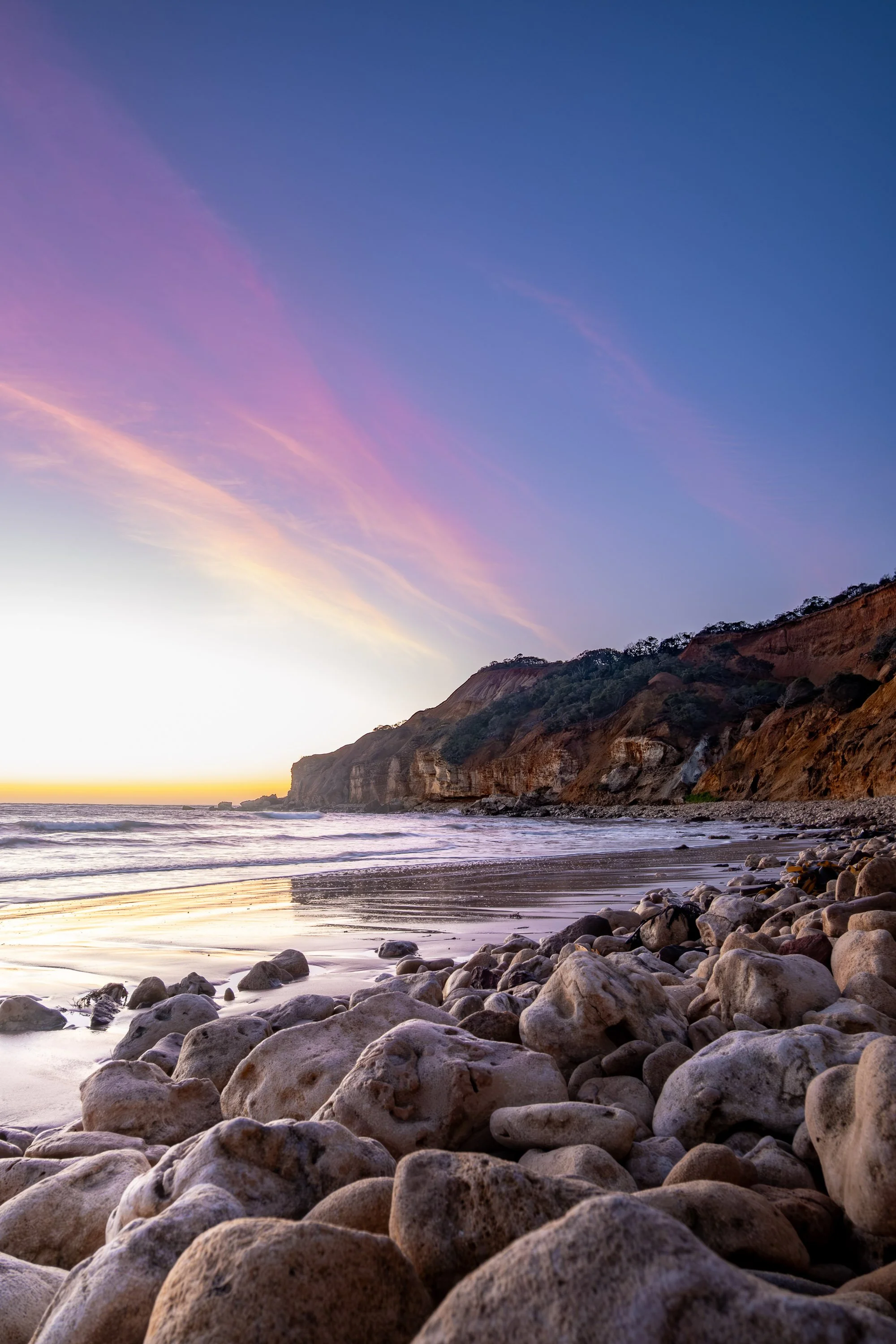 Scenic view of a rocky beach at sunset with cliffs in the background and a sky with pastel pink, purple, and blue clouds.