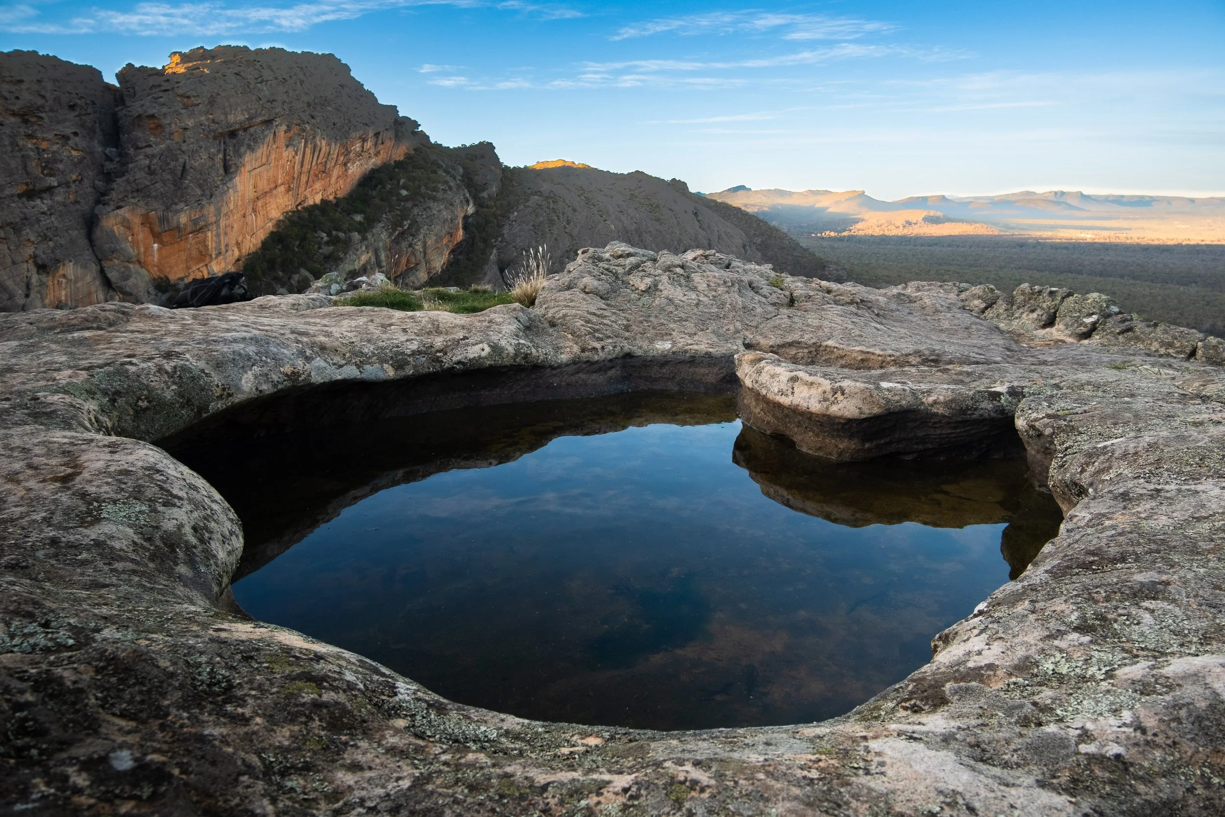 A small, natural rock pool filled with water on a flat rocky surface, overlooking a vast mountainous landscape and clear sky.