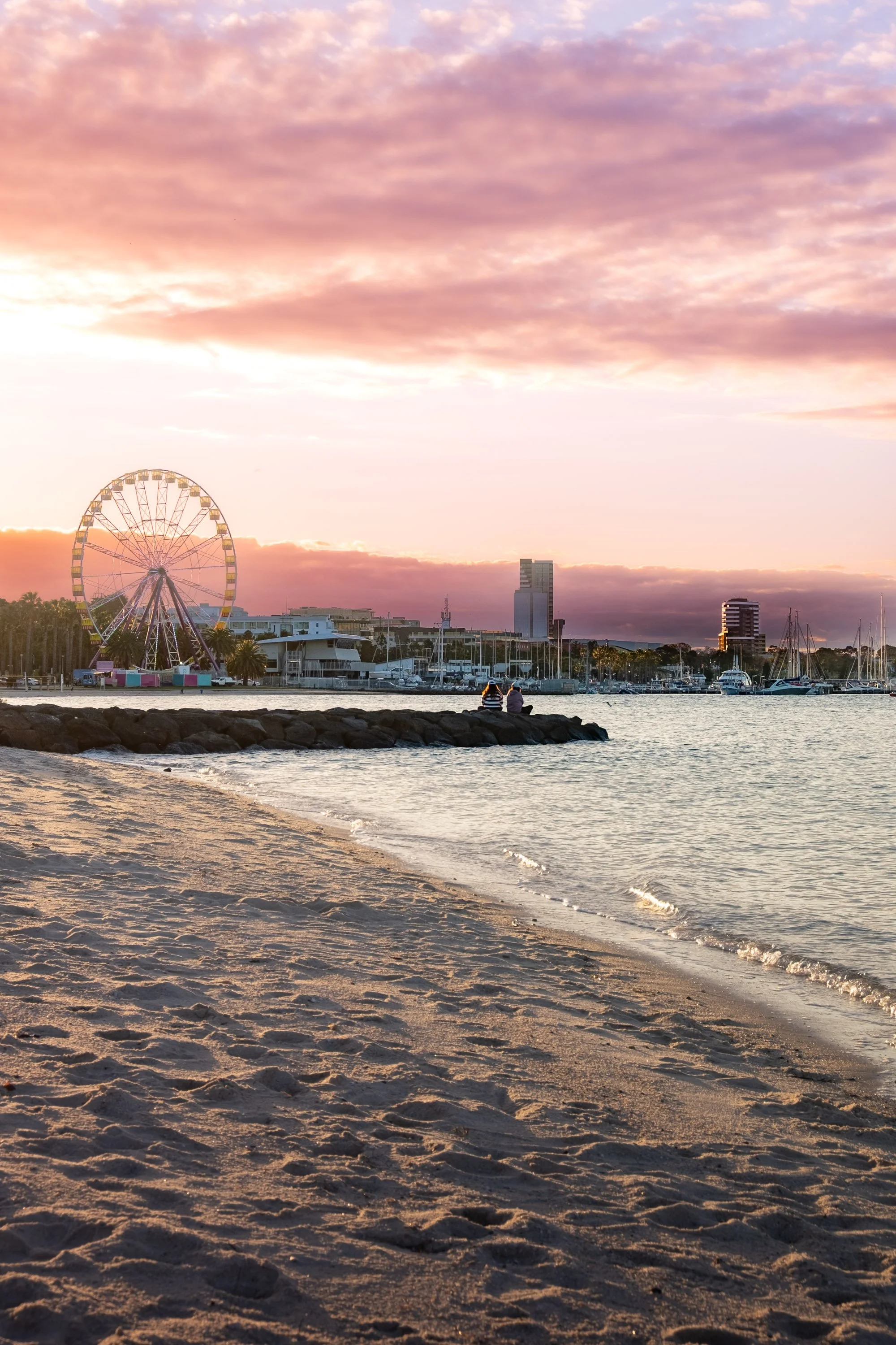 Sunset over a sandy beach with a ferris wheel, boats in the harbor, and two people sitting on a rocky jetty