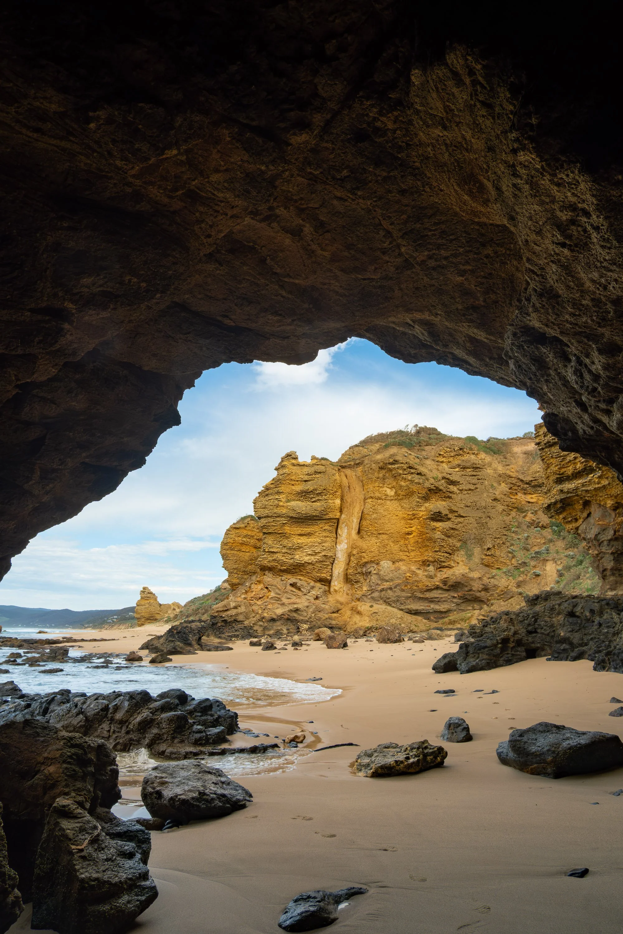 View of a sandy beach through a rocky cave opening, with large rocks and cliffs along the shoreline and a blue sky with some clouds overhead.
