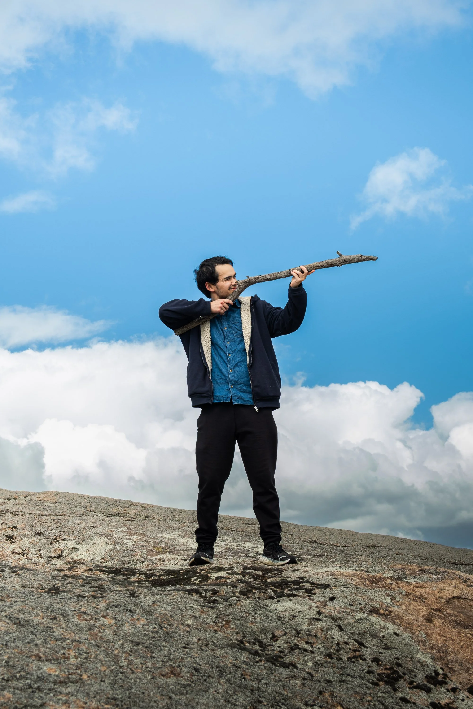 A man standing on a rocky terrain holding a large stick up to his mouth as if he is blowing a horn, with a blue sky and clouds in the background.