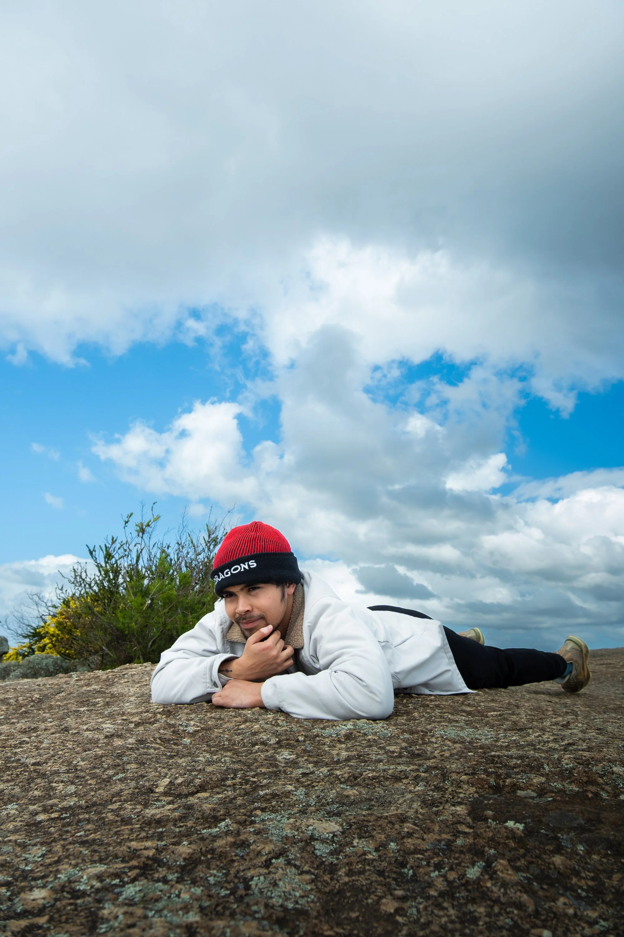 A man lying on a large rock outdoors under a cloudy sky, wearing a red and black beanie, a light jacket, and black pants.