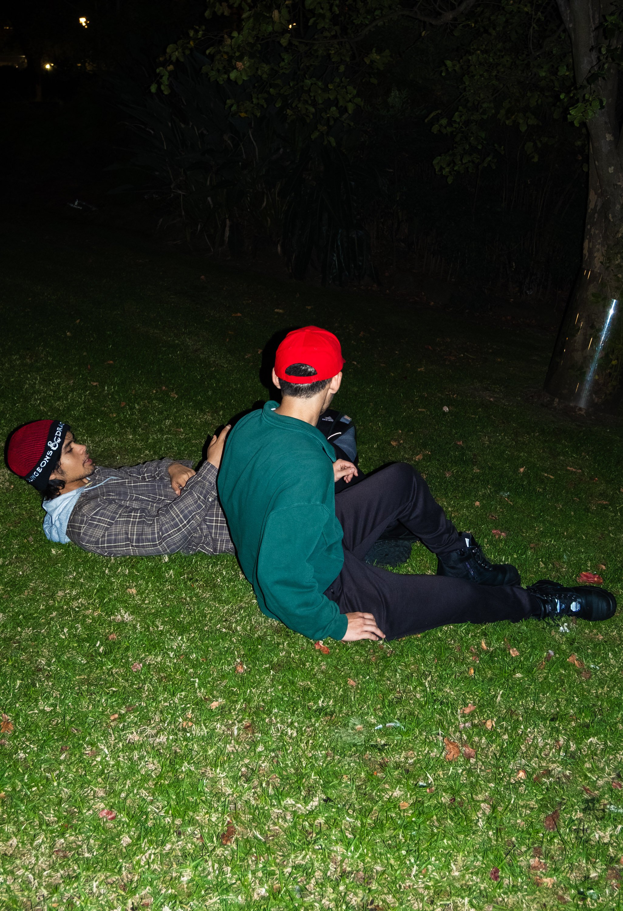 Two young men sitting on grass at night, one lying down and the other sitting with one hand on the ground, near trees and a pole.