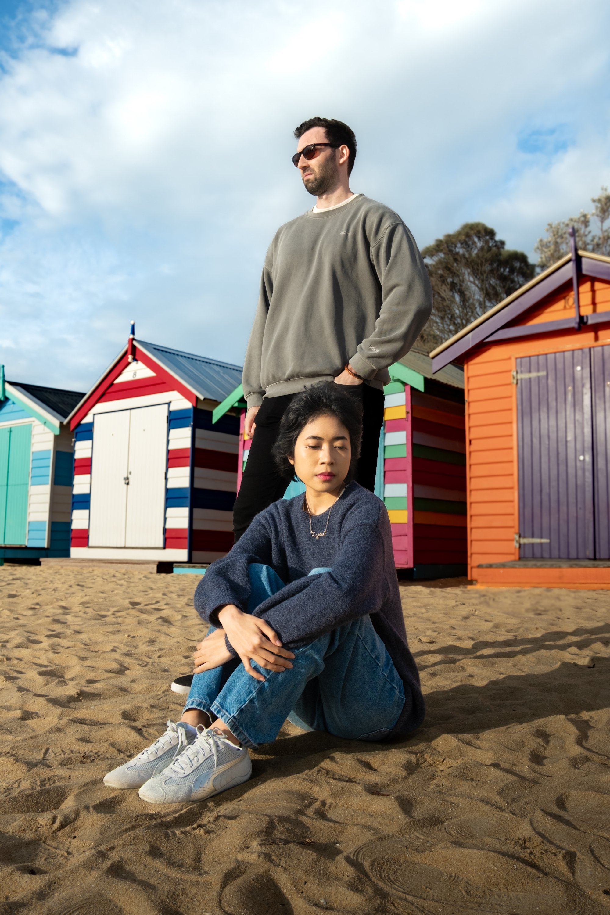 A man and woman on a sandy beach with colorful beach huts behind them. The woman sits cross-legged on the ground with eyes closed, wearing a dark sweater, jeans, and sneakers. The man stands behind her with hands in pockets, wearing a gray sweatshirt