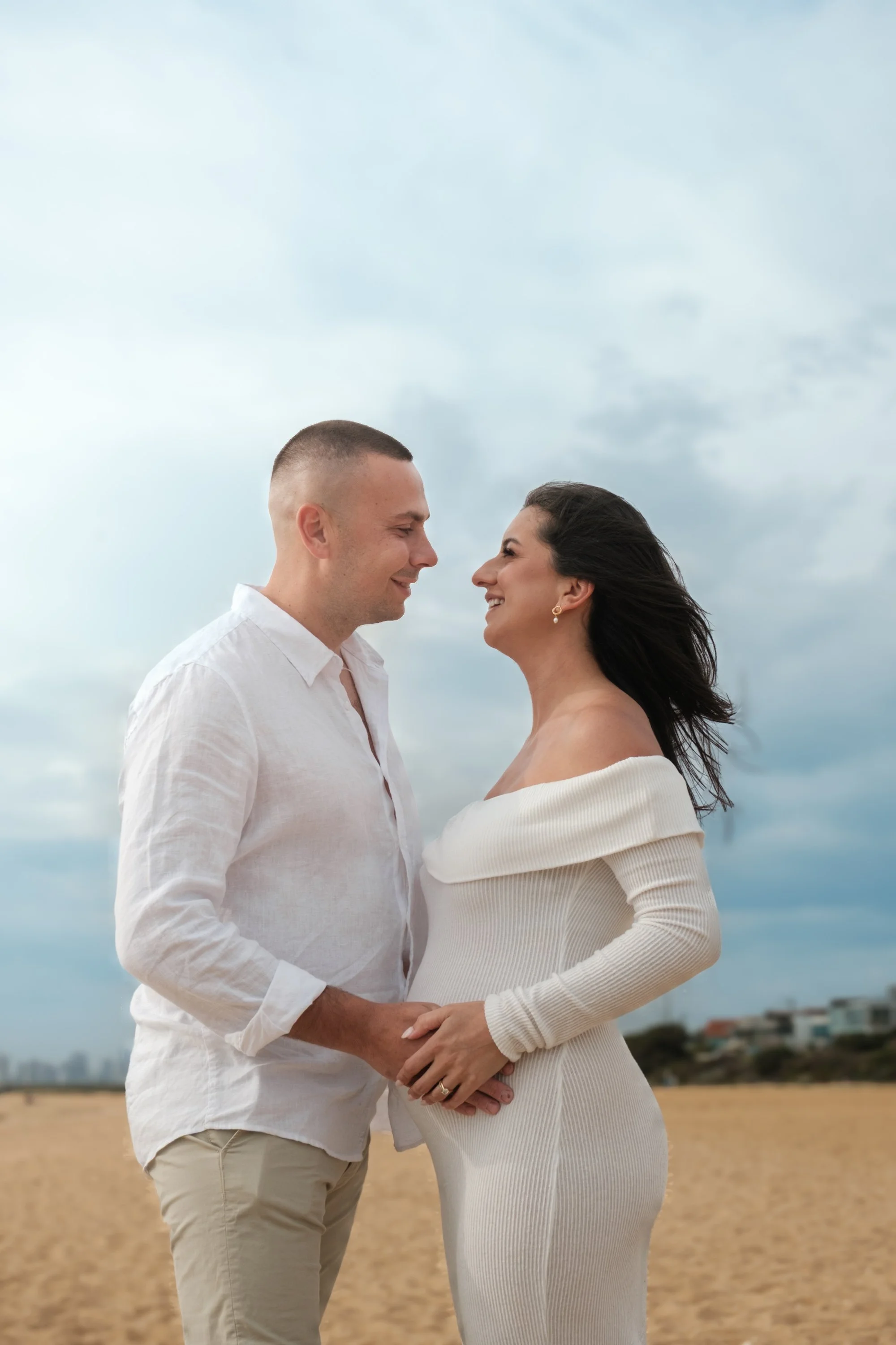 A couple holding hands and smiling at each other on a beach with a cloudy sky in the background.
