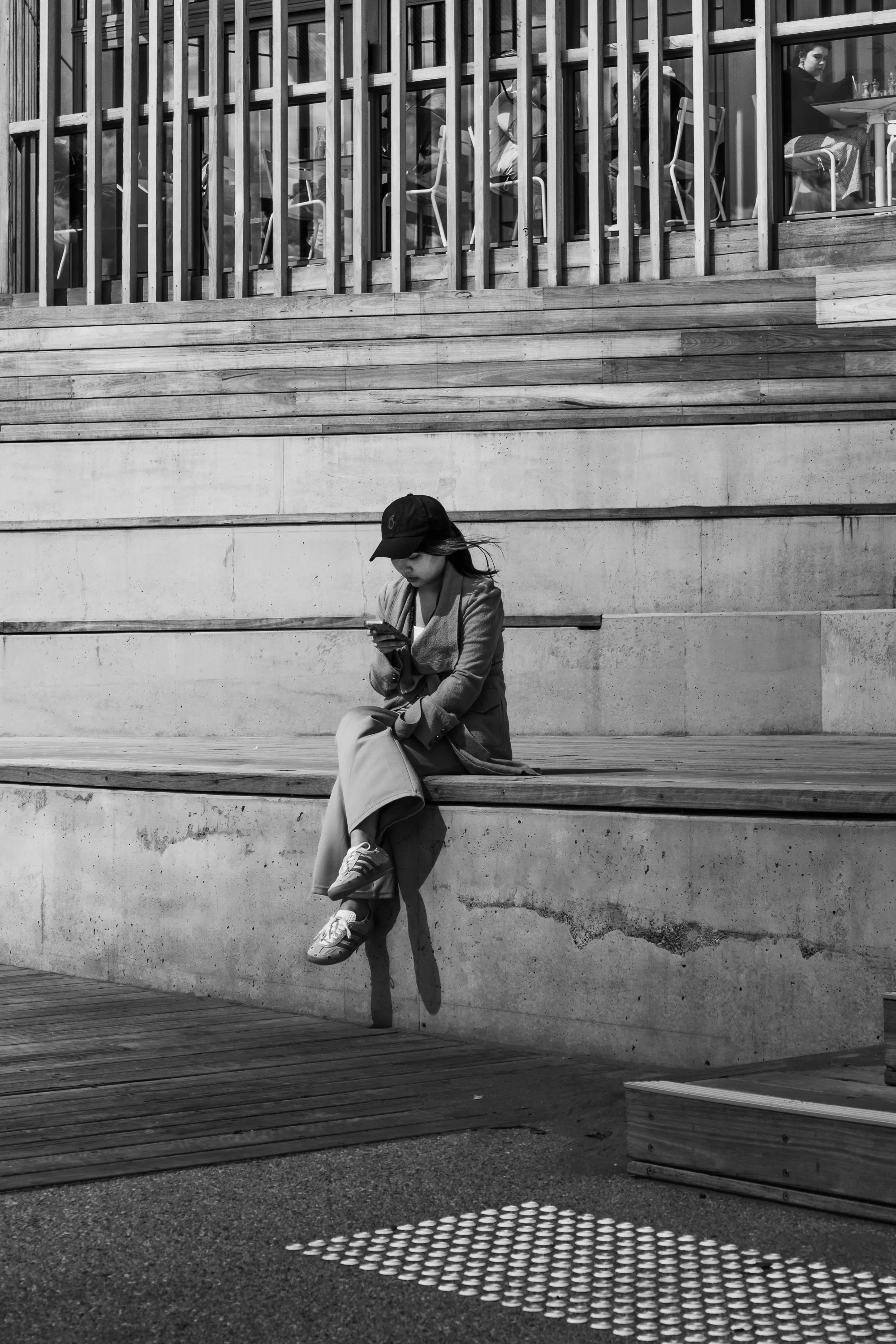 A woman sitting on a concrete bench, looking at her phone, outdoors with a modern wooden and glass building behind her, in black and white.