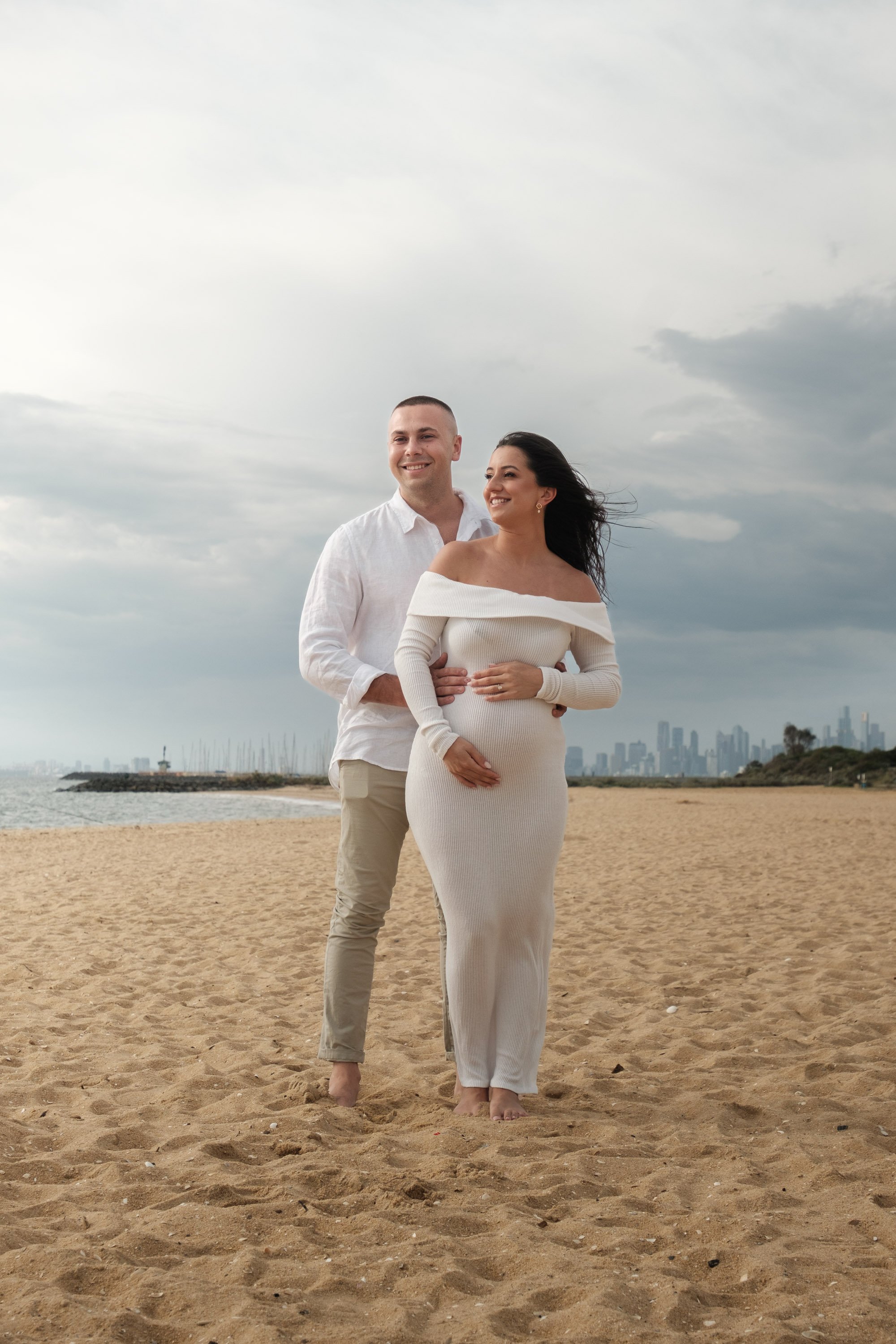 A happy couple, with the woman visibly pregnant, walking on a sandy beach with a city skyline in the background on a cloudy day.