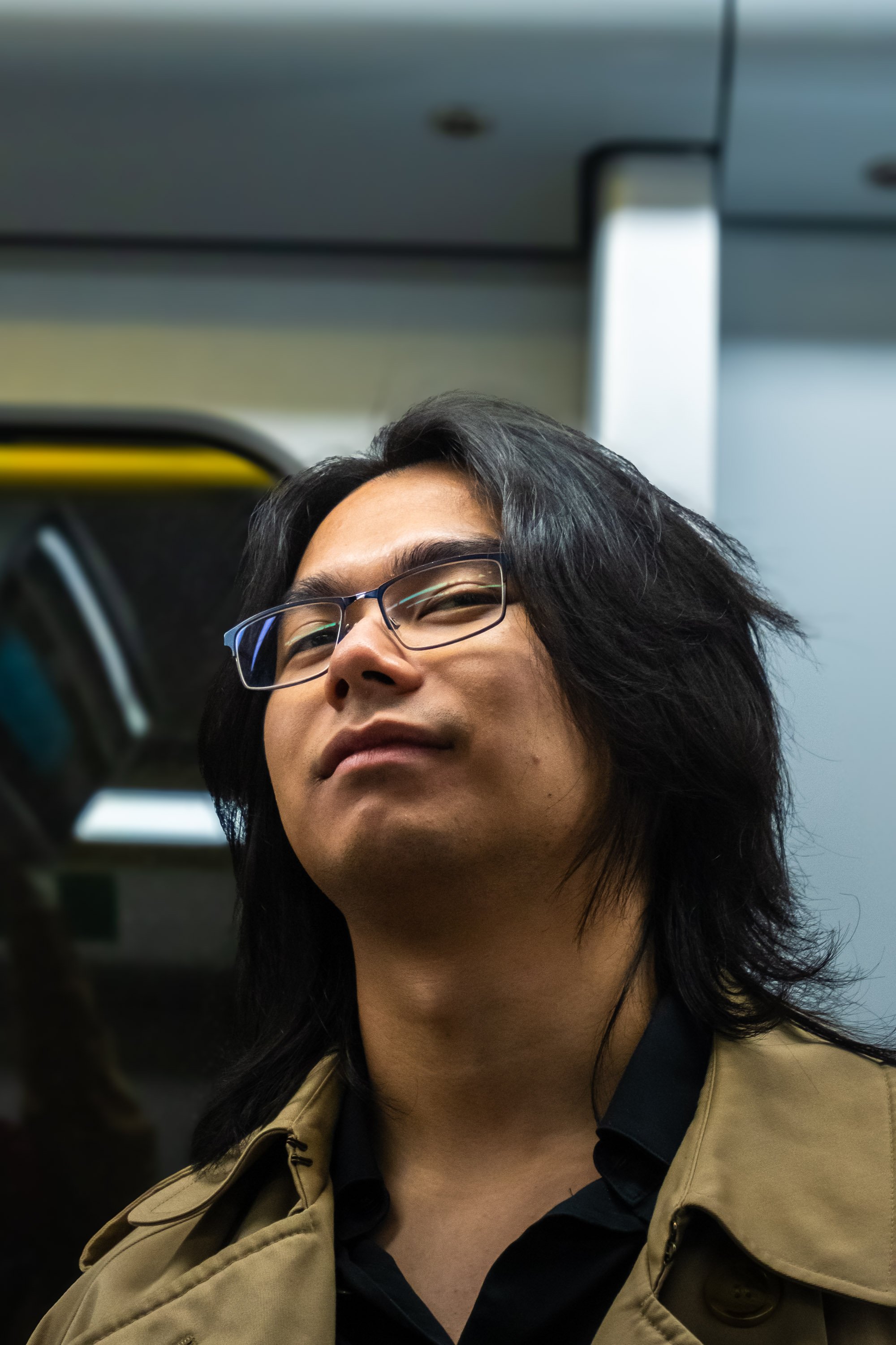 Close-up of a man with long black hair and glasses, wearing a beige coat and black shirt, standing in a subway or train station.