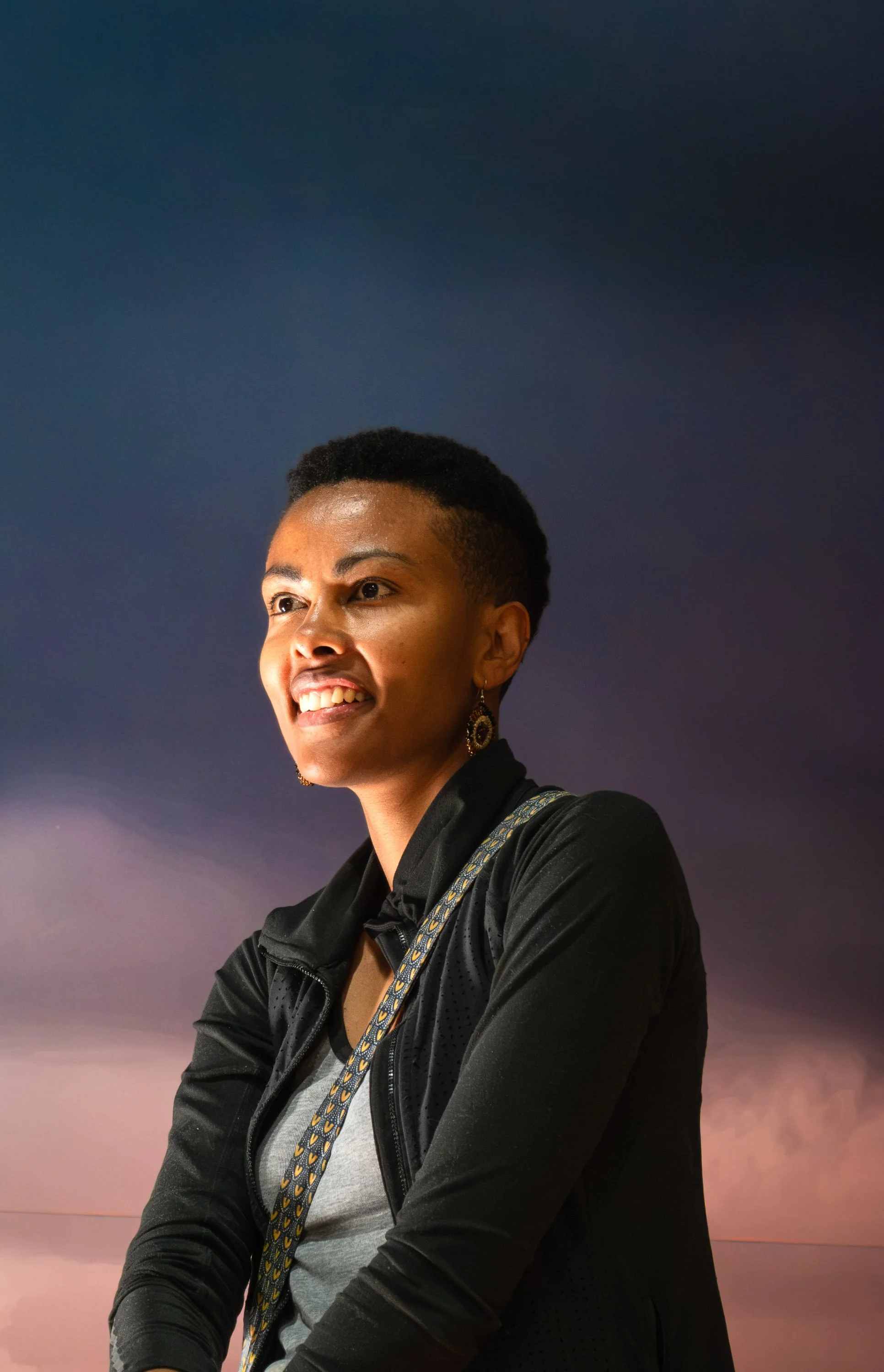A woman with short hair, wearing a black jacket, earrings, and a backpack strap, stands outdoors at sunset or dusk with a colorful sky in the background.