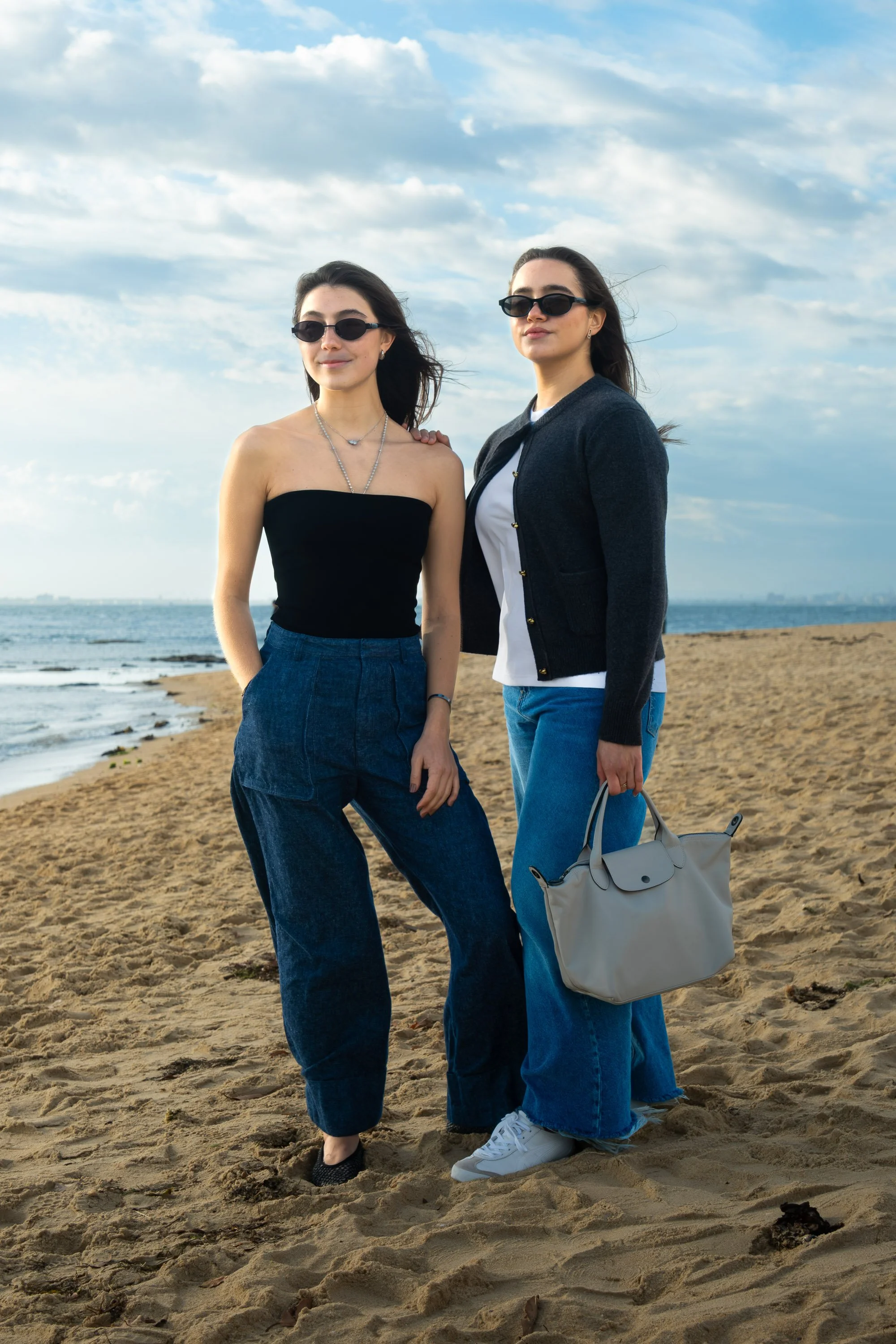Two women standing on a sandy beach under cloudy sky, wearing sunglasses and casual clothing, with the ocean in the background.