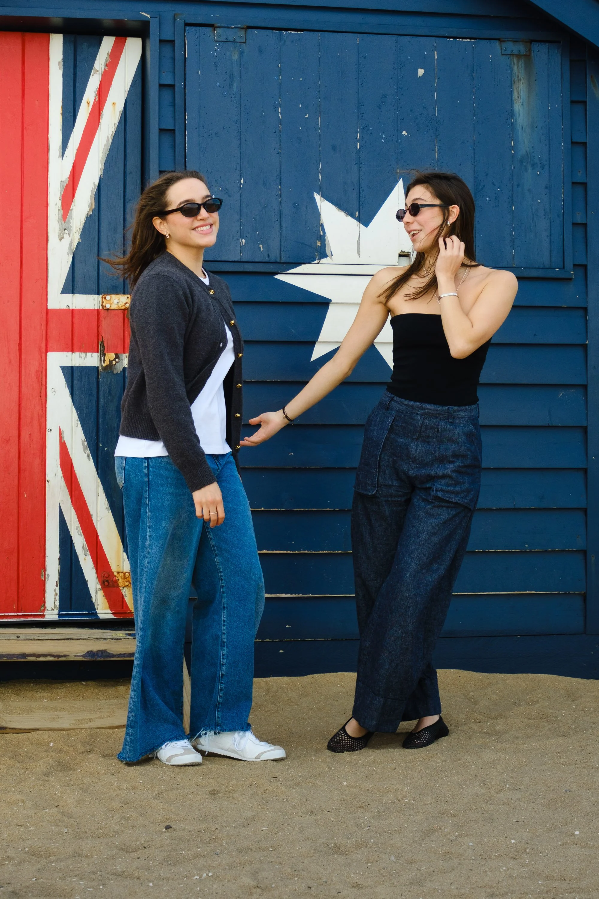 Two women standing outdoor near a building painted with a stylized Union Jack flag, both wearing sunglasses and casual clothing, smiling and interacting with each other.