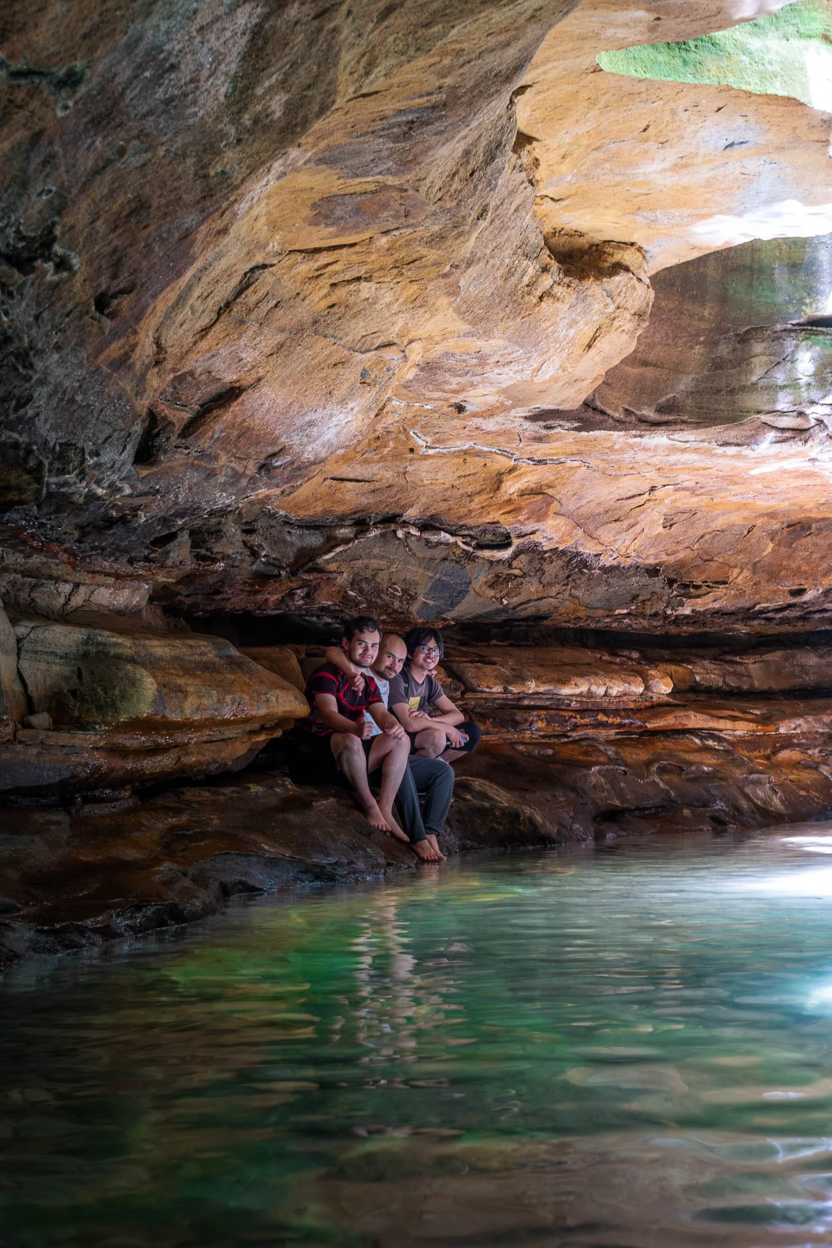 Three friends sitting on rocks by water inside a cave with a large rock ceiling overhead.