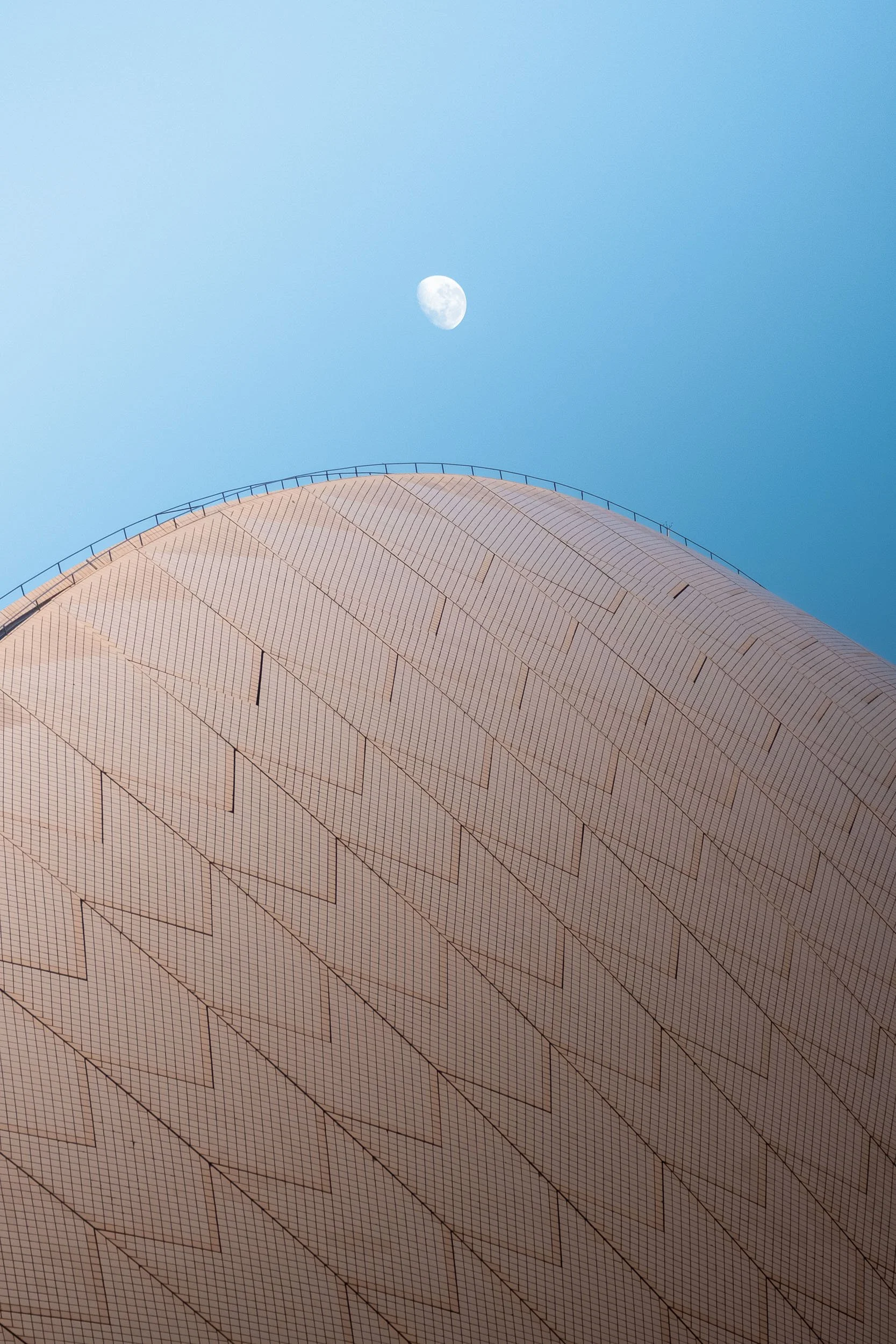 Close-up of a large modern building with a rounded, tiled facade against a clear blue sky, with the moon visible in the background.