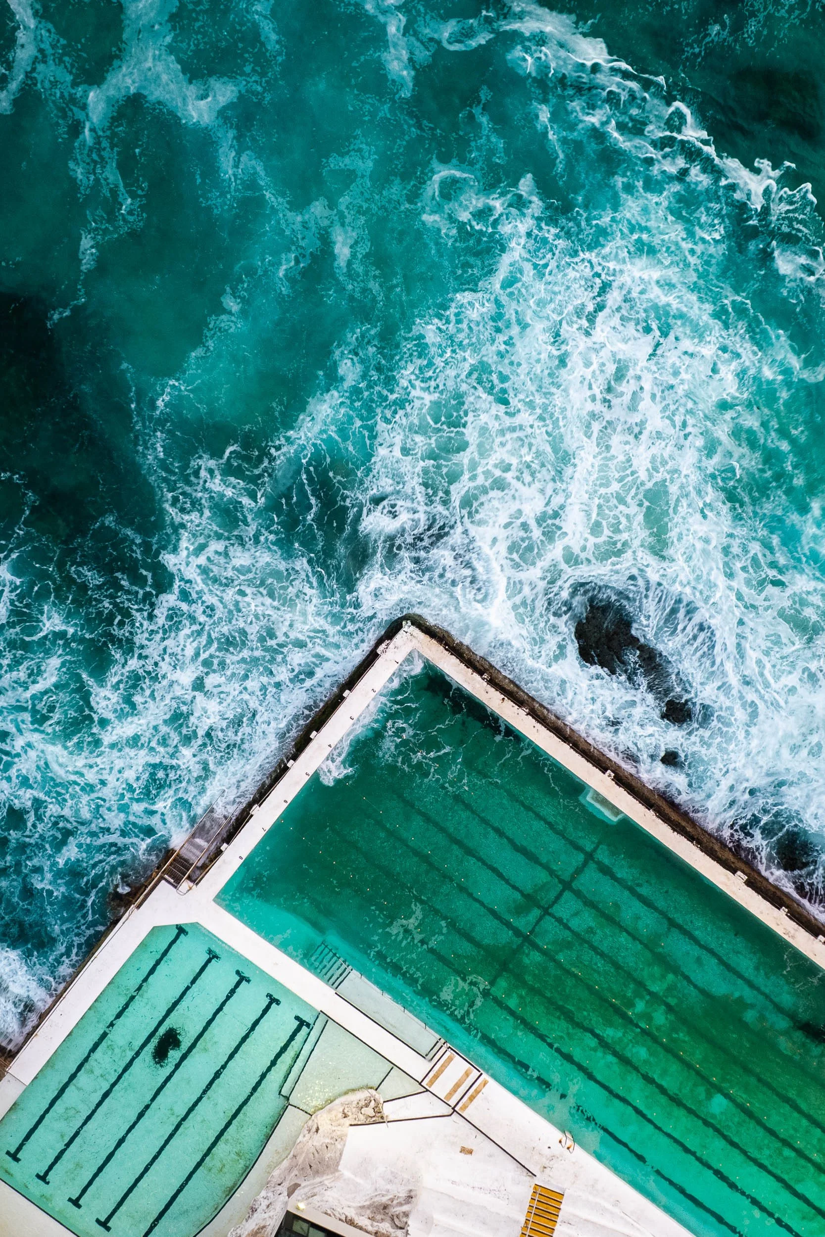 An aerial view of a swimming pool next to rough ocean waves crashing over a rocky barrier.