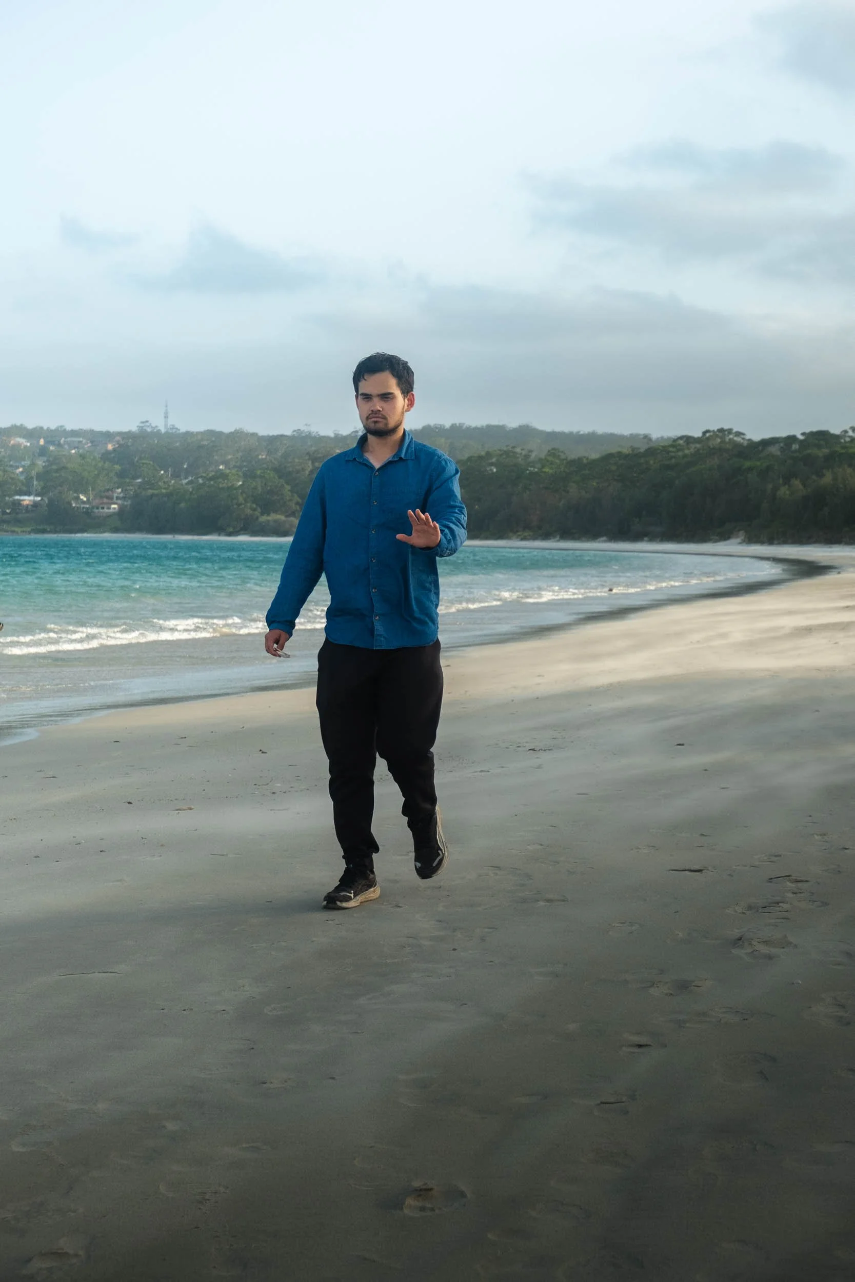 A man walking on a sandy beach near the ocean, with trees and a cloudy sky in the background.