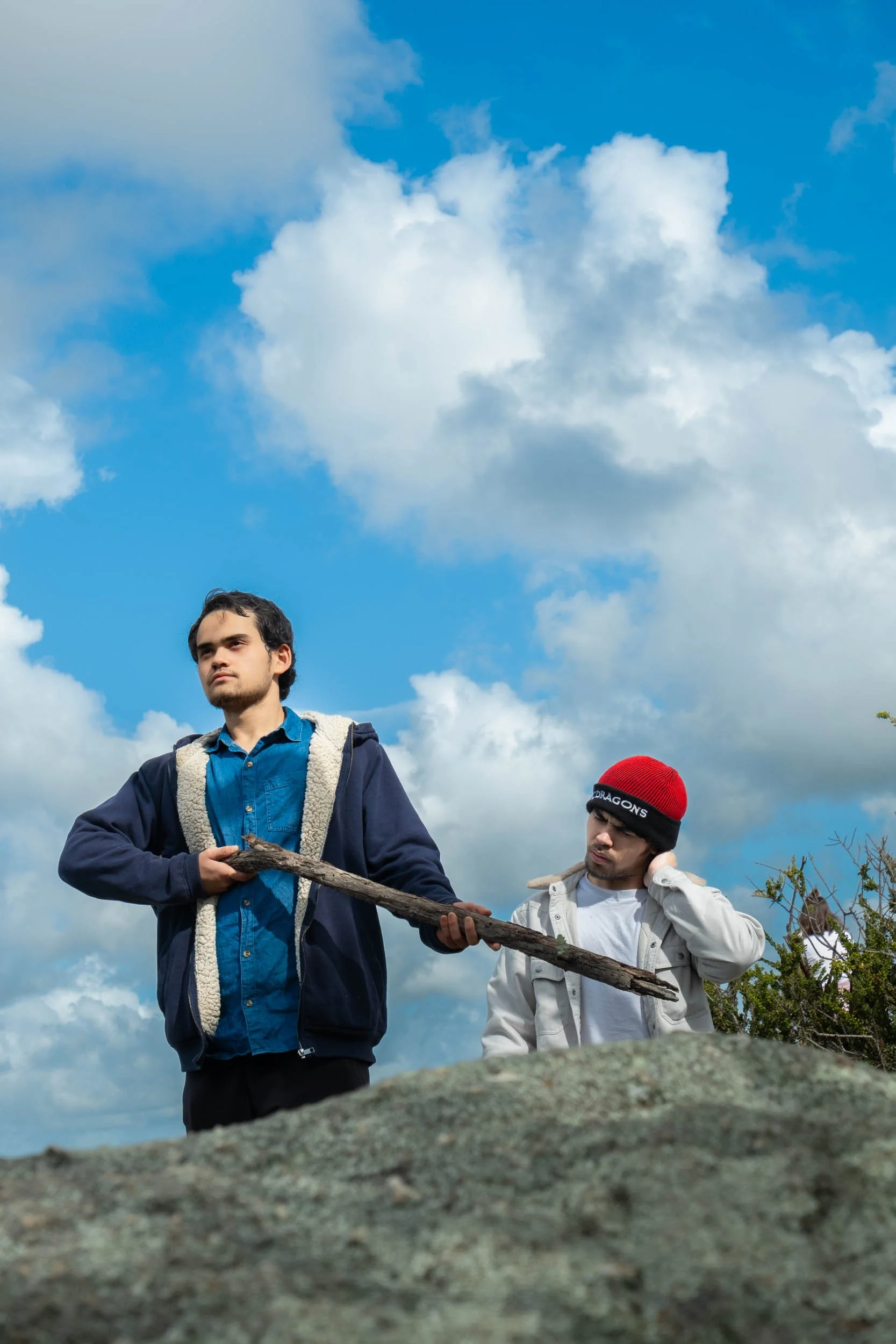 Two young men outdoors against a blue sky with white clouds. One is holding a long stick, and the other appears to be adjusting his hat.