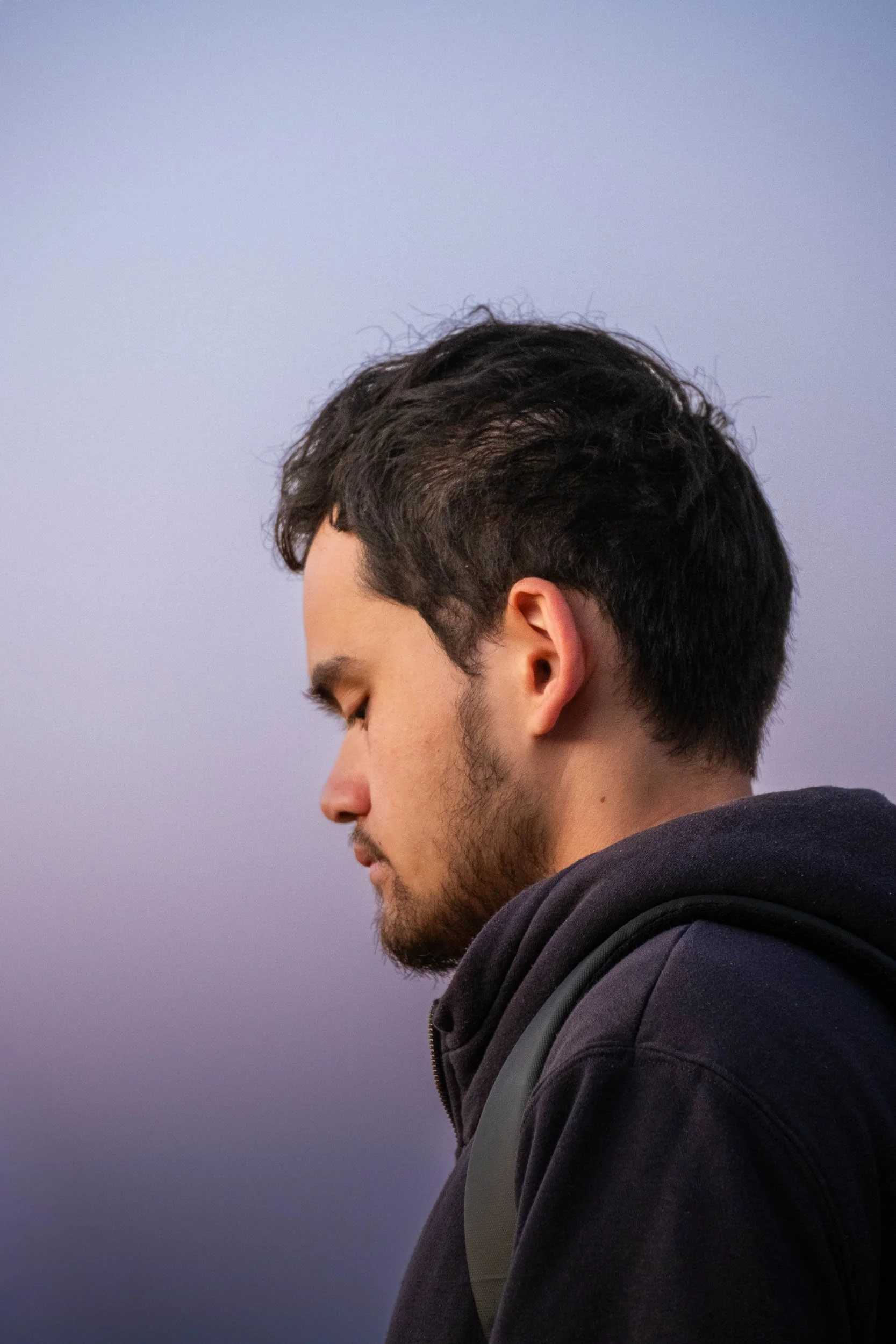 Side profile of a young man with dark hair and beard, wearing a dark hoodie, against a blurred purple sky.