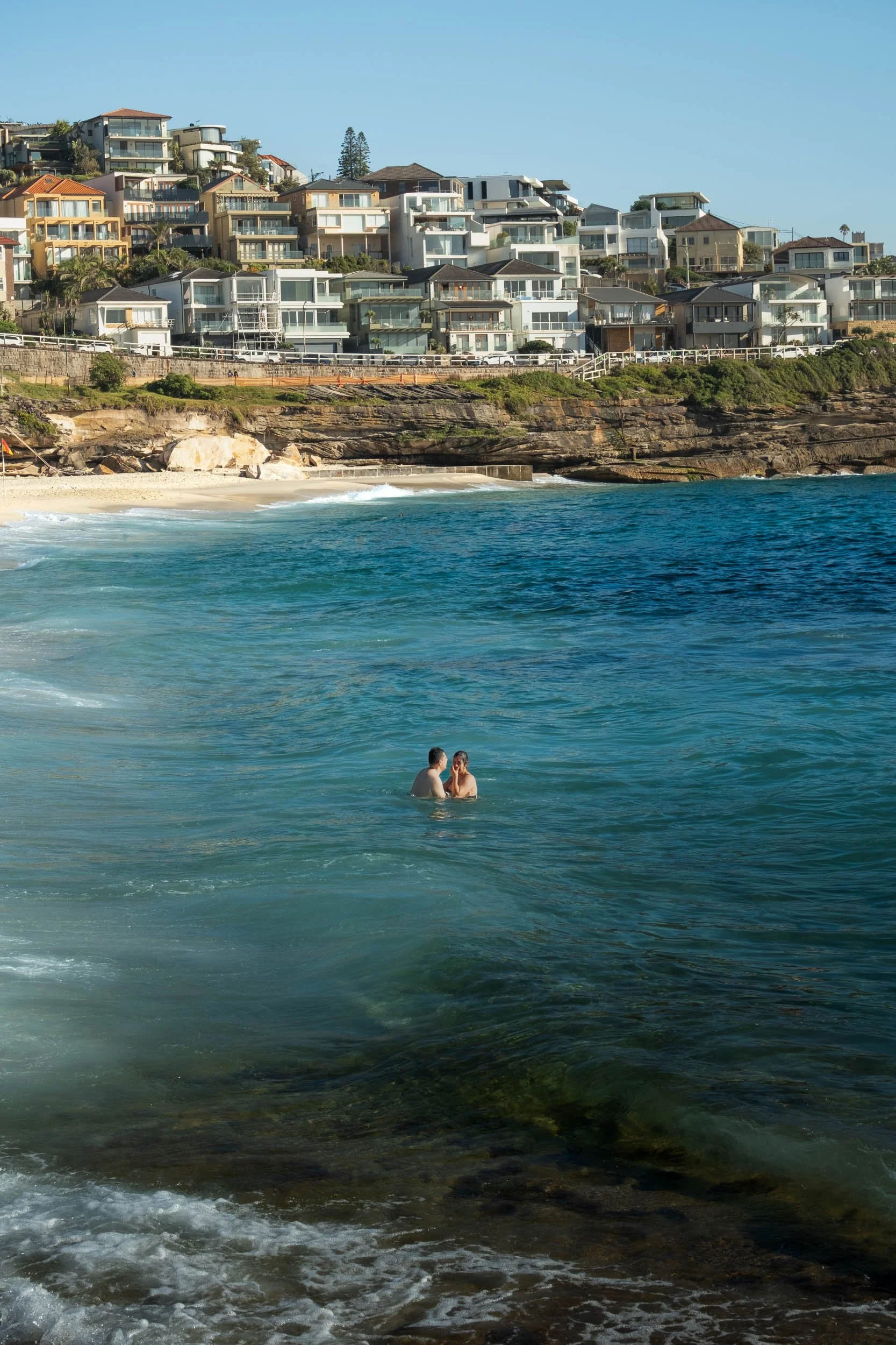 A couple swimming in the ocean near a sandy beach with residential houses on a hill in the background.