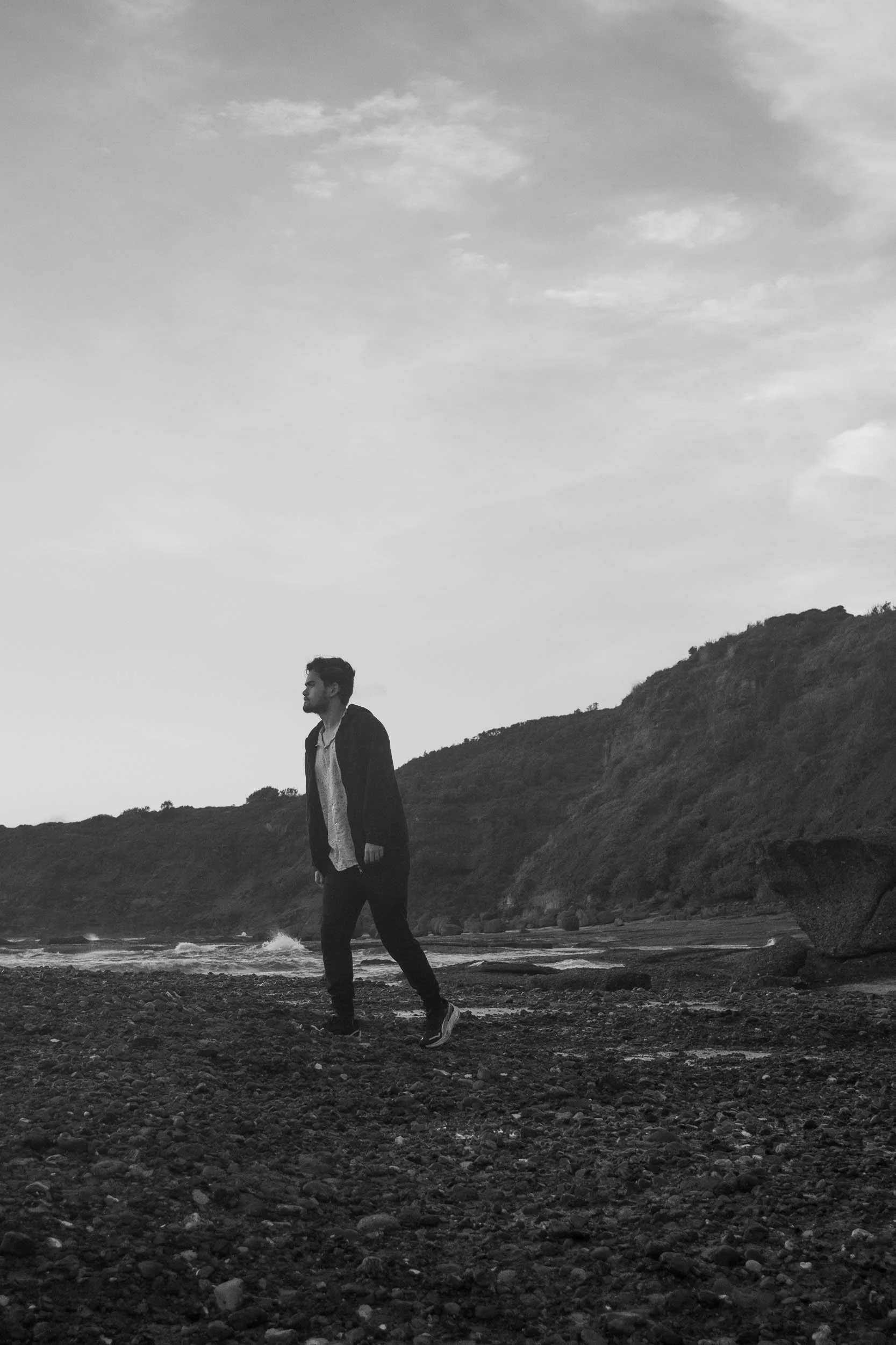 A man walking on a rocky beach near cliffs, in black and white.