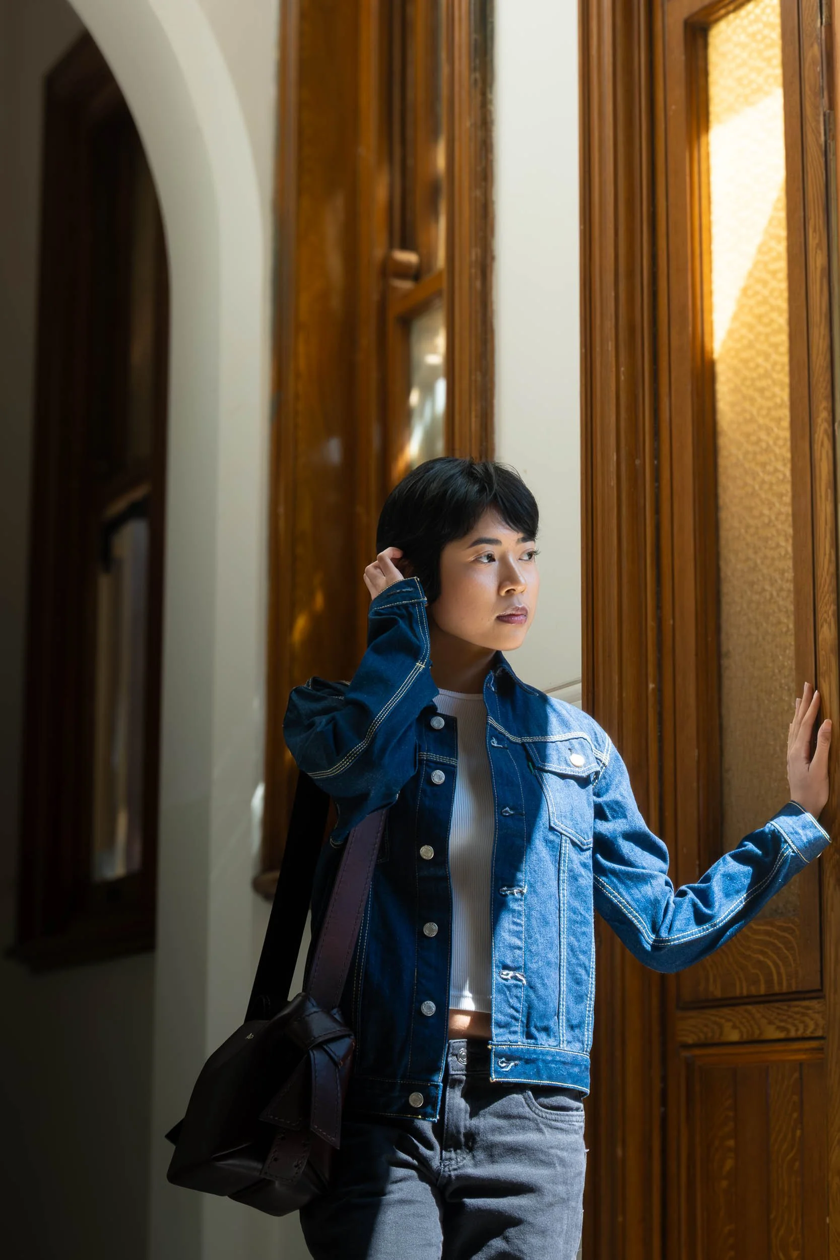 Young woman with short black hair, wearing a denim jacket, standing near a wooden door in an indoor setting, holding the door with one hand and touching her ear with the other.