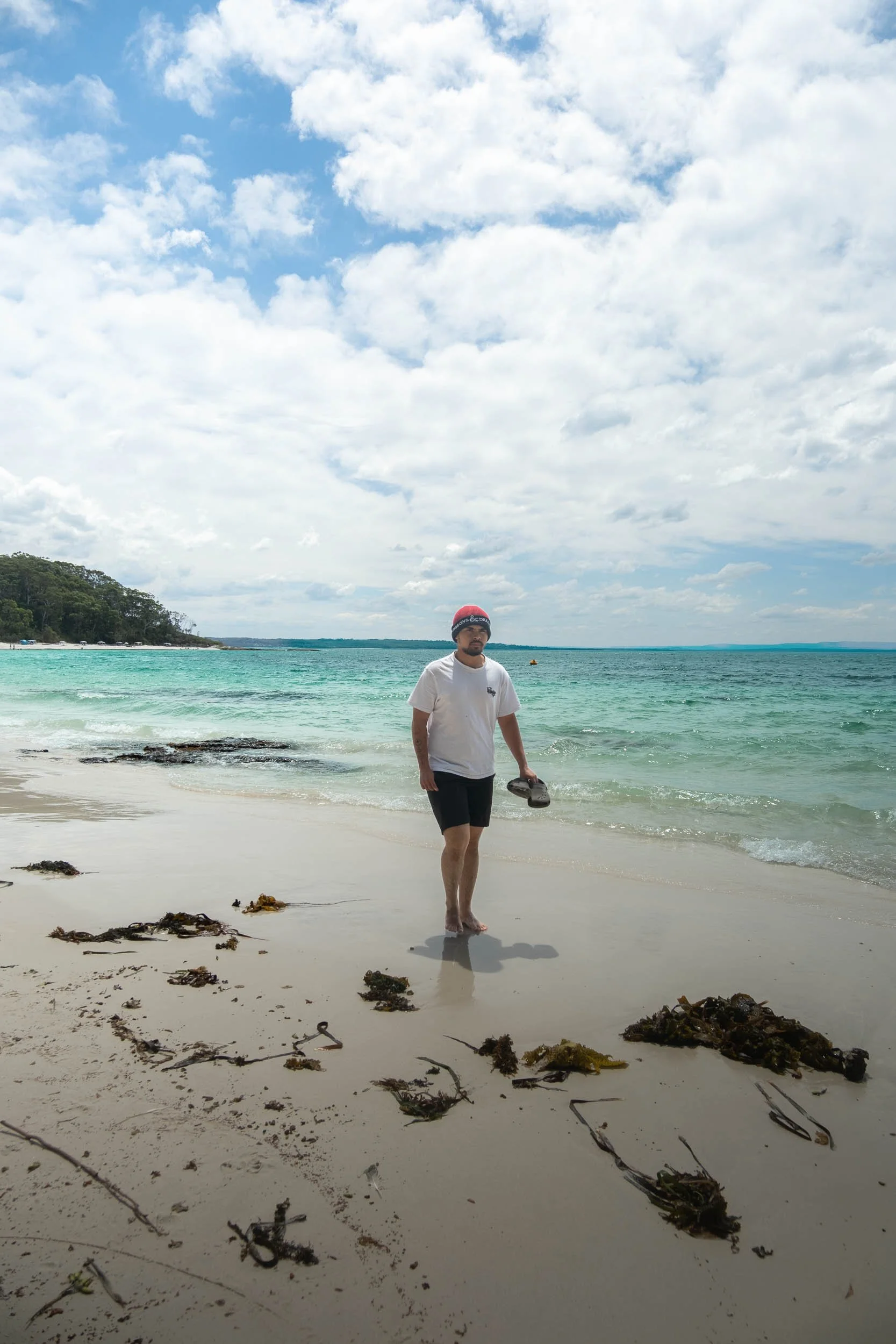 Man walking barefoot on beach holding pair of shoes, wearing a white t-shirt, black shorts, and a red beanie, with seaweed scattered on white sand, ocean in the background, cloudy sky above.