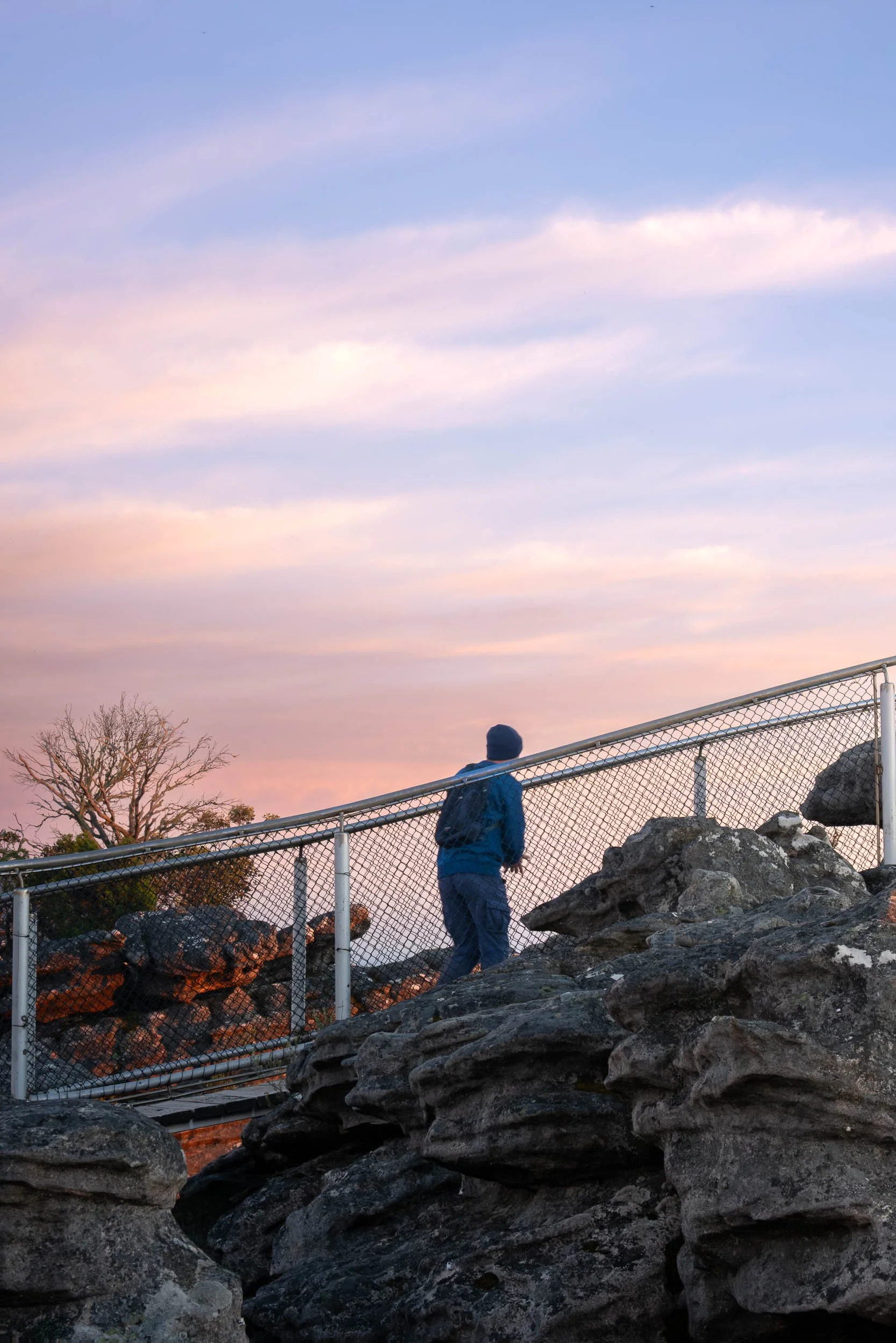 A person wearing a blue jacket, jeans, and a beanie, walking on a rock pathway near a chain-link fence during sunset.