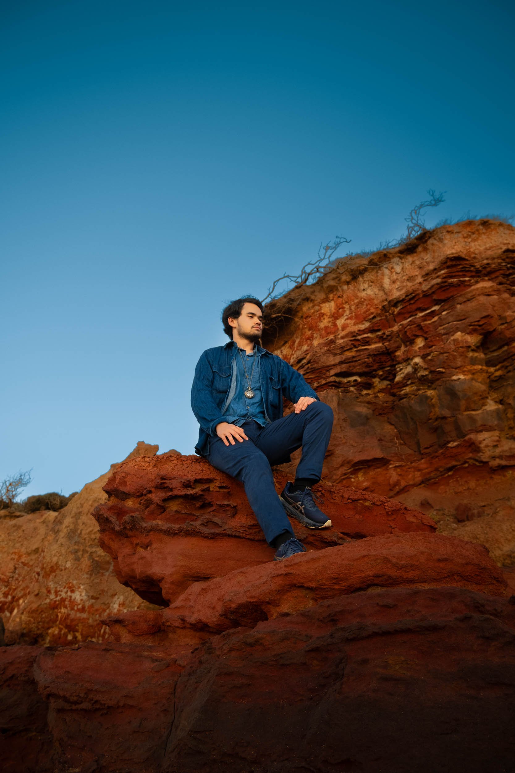 A young man in blue jeans, a denim shirt, and dark sneakers, sitting on a large reddish rock formation in a desert landscape with clear blue sky.