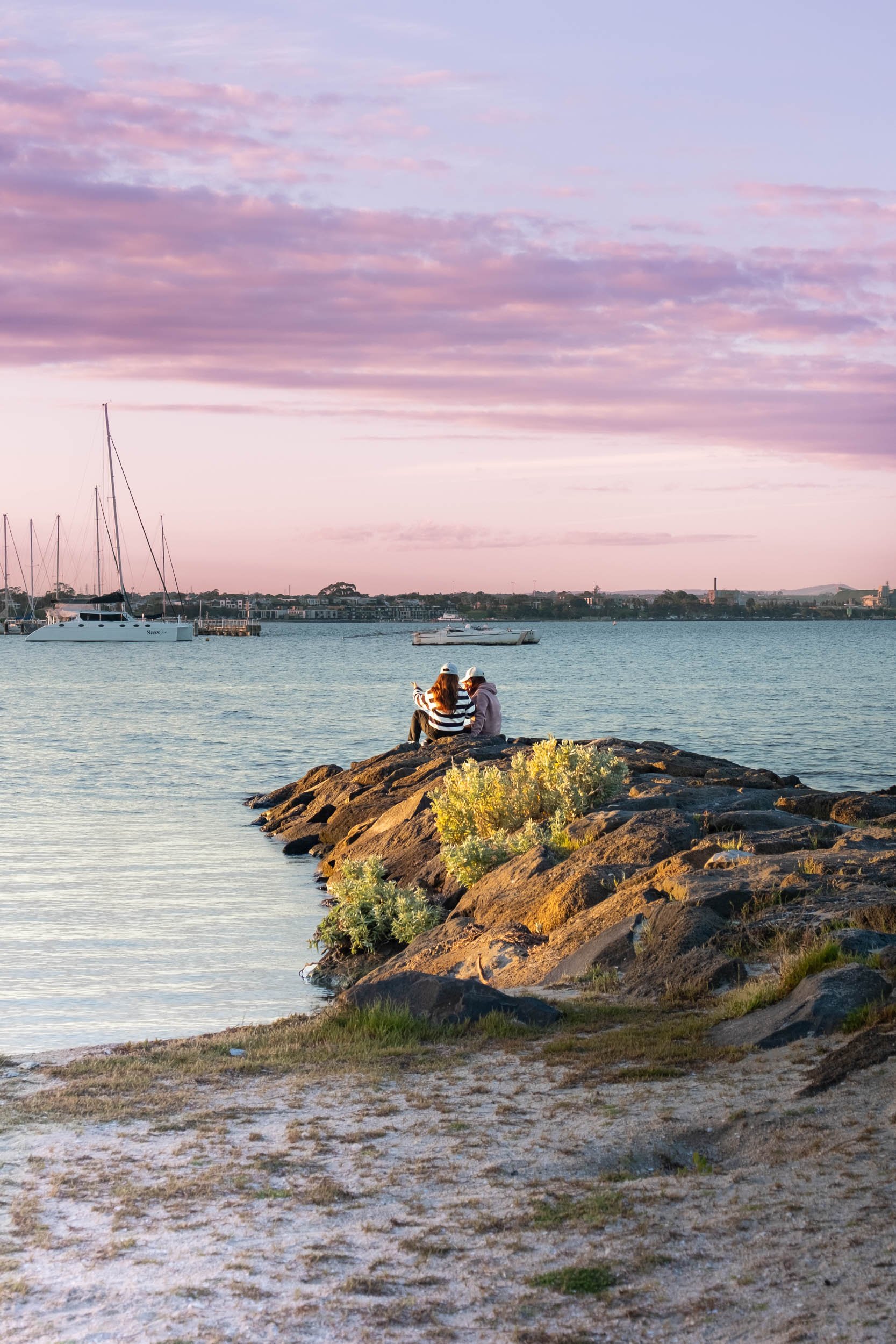 Two women sit on rocks by the water at sunset, with boats anchored in the background and lavender-colored clouds in the sky.