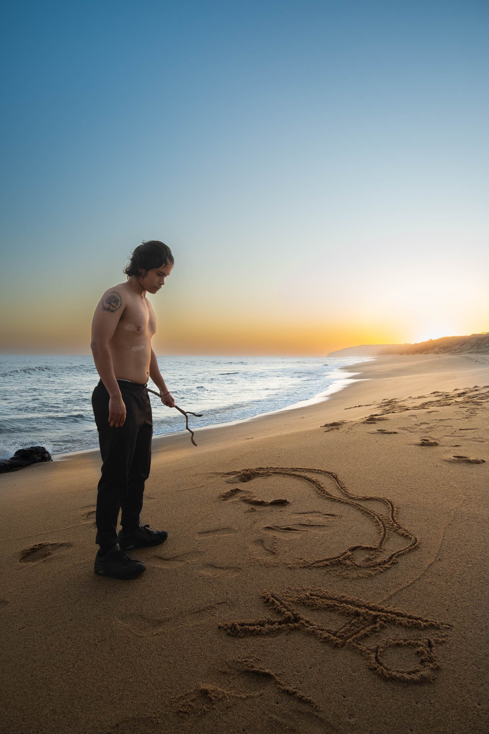 A man stands on a sandy beach during sunset, looking at a drawing of a whale and a fishing hook in the sand.