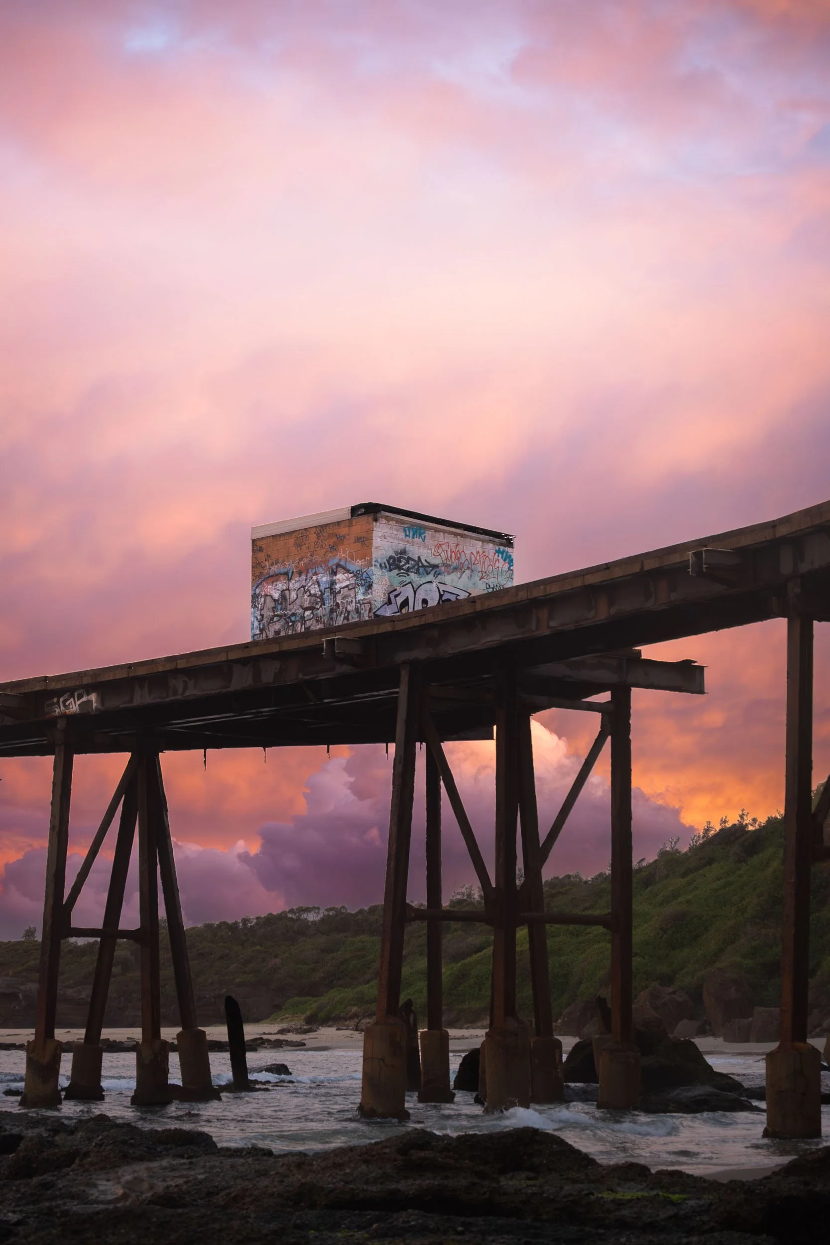 An old wooden pier over the ocean with a graffitied small building on top, set against a colorful sunset sky with pink and purple clouds.
