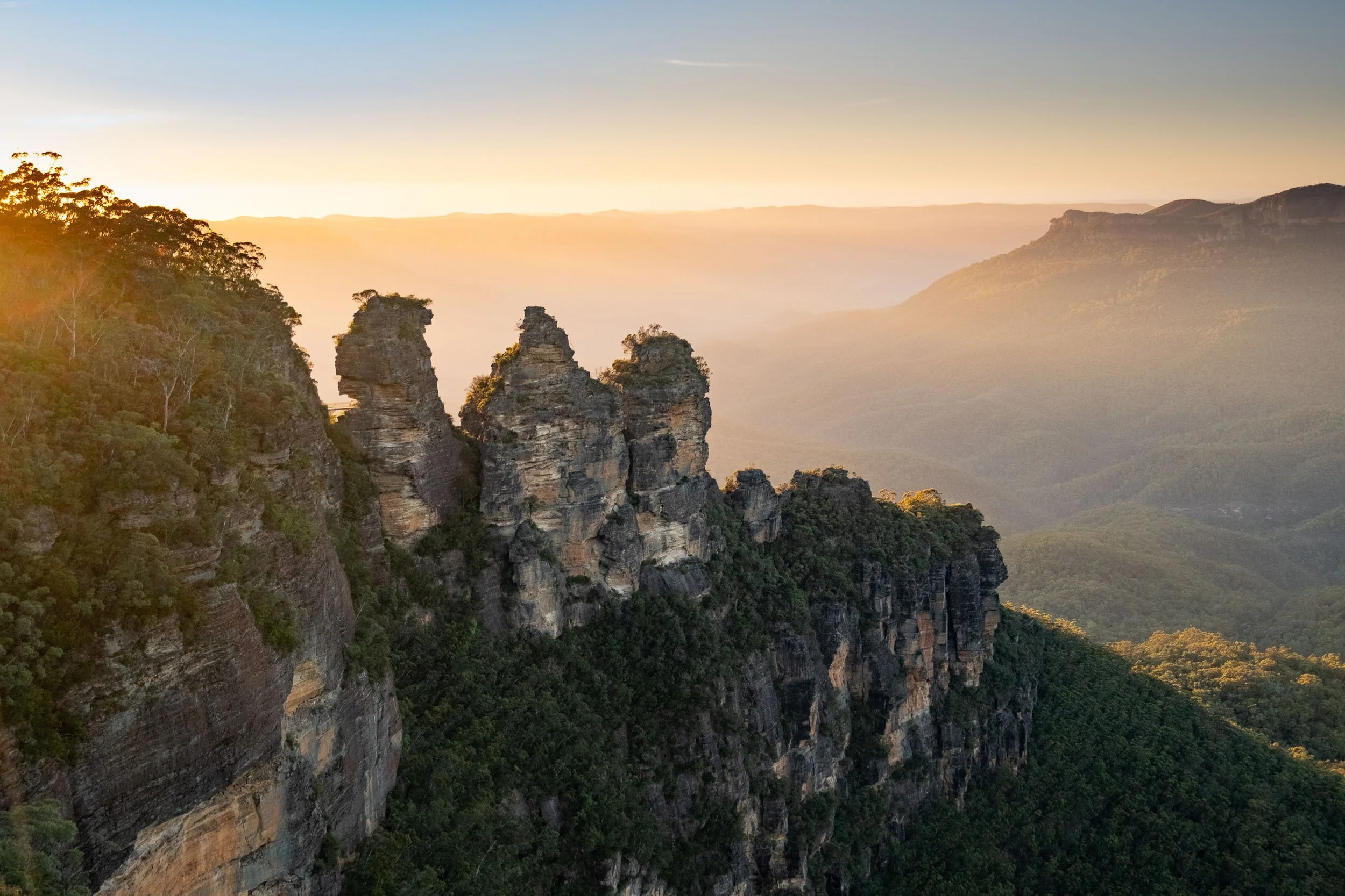 Sunrise over the Three Sisters rock formation in the Blue Mountains, surrounded by lush green trees and distant mountain ranges.