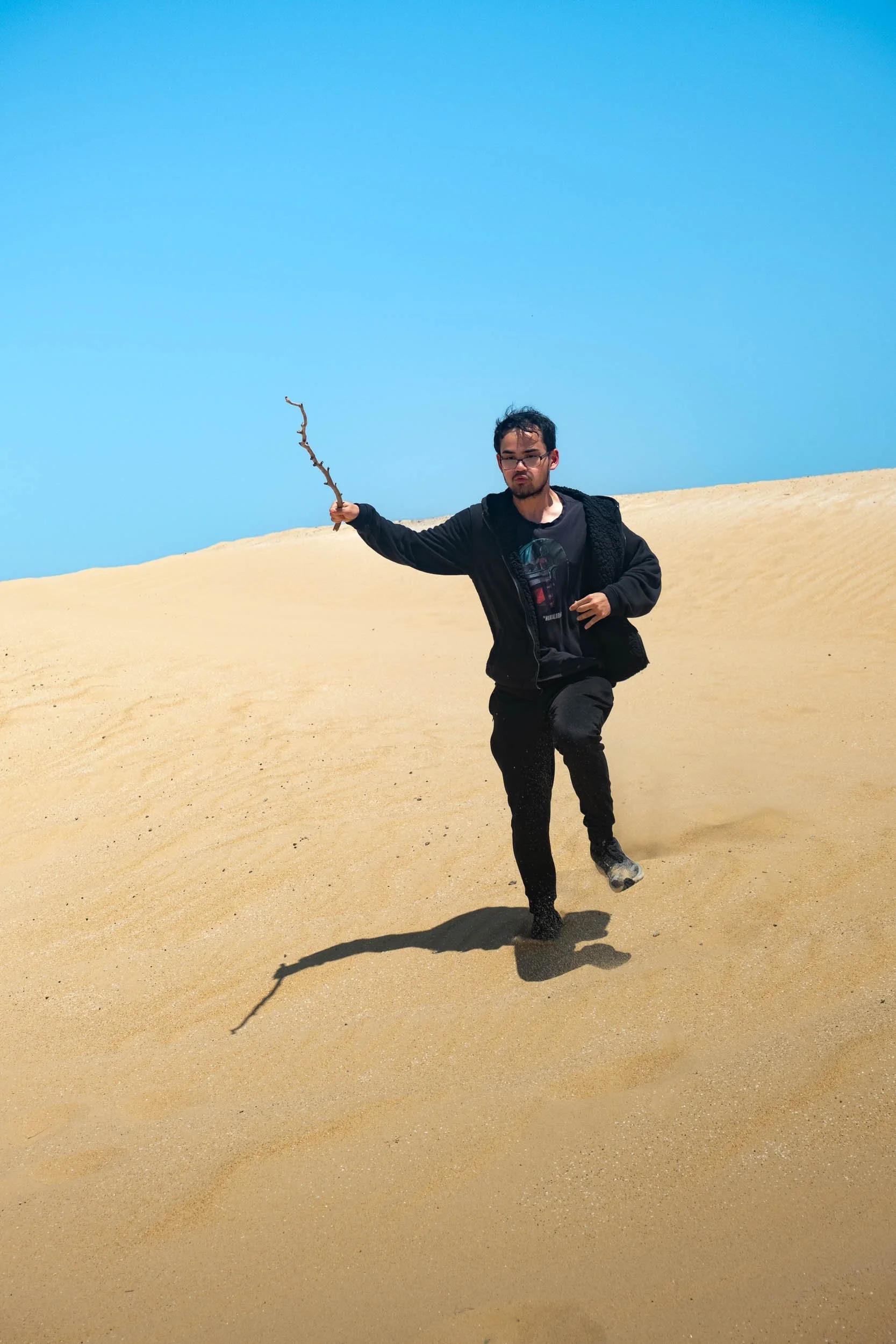 A man running on sand dunes under a clear blue sky, holding a small branch.