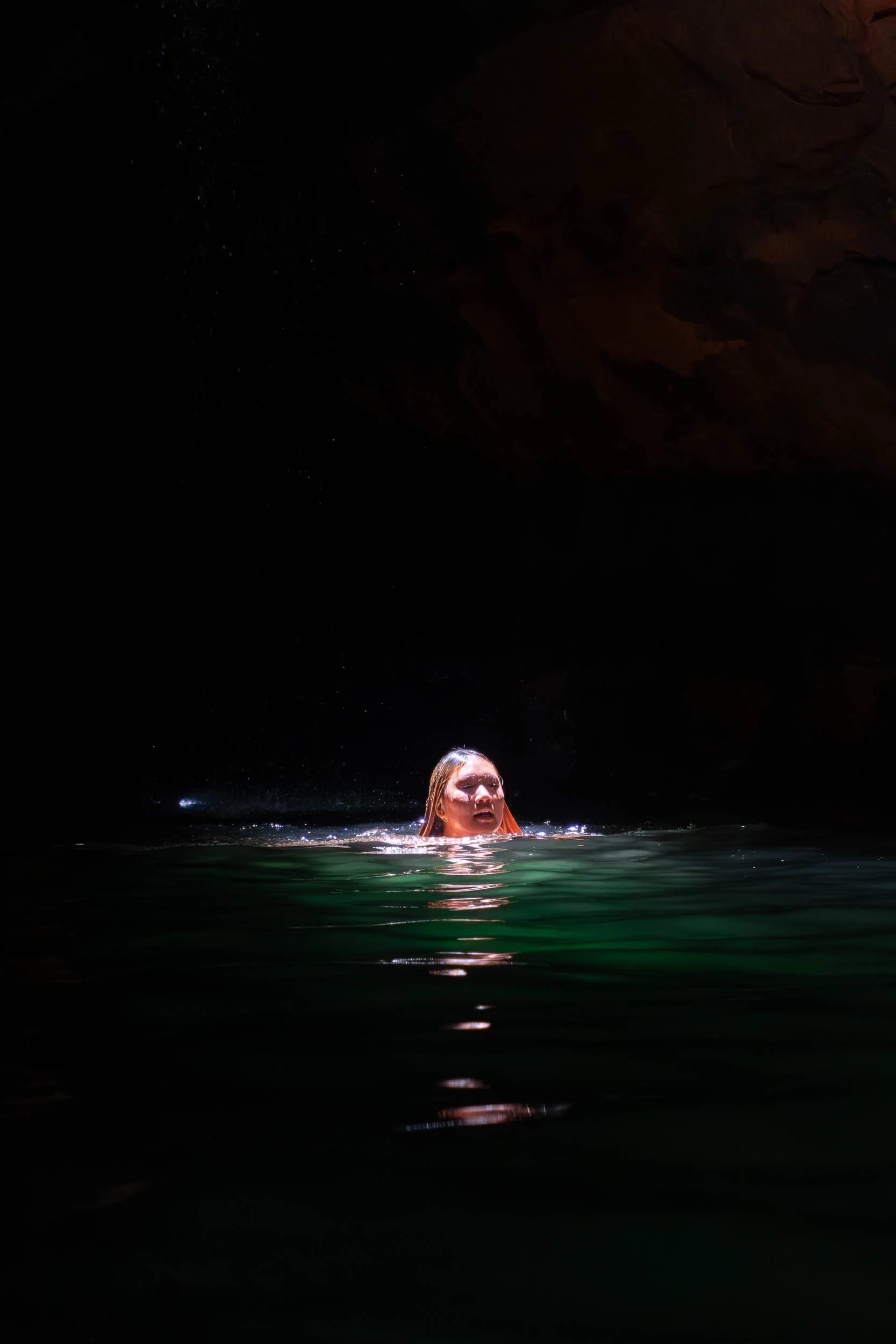 A woman swimming in a dark cave with water reaching her shoulders, illuminated by a light source above.