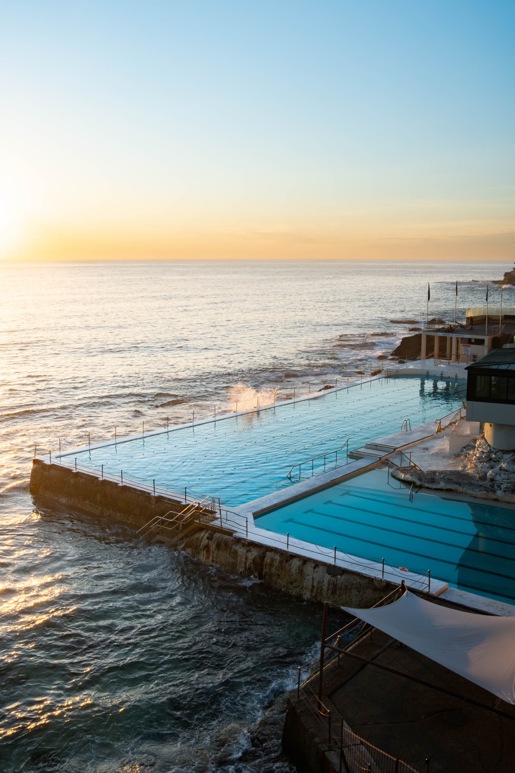 Outdoor pool with ocean view at sunset, waves crashing against the pool structure, umbrellas and lounge chairs nearby.
