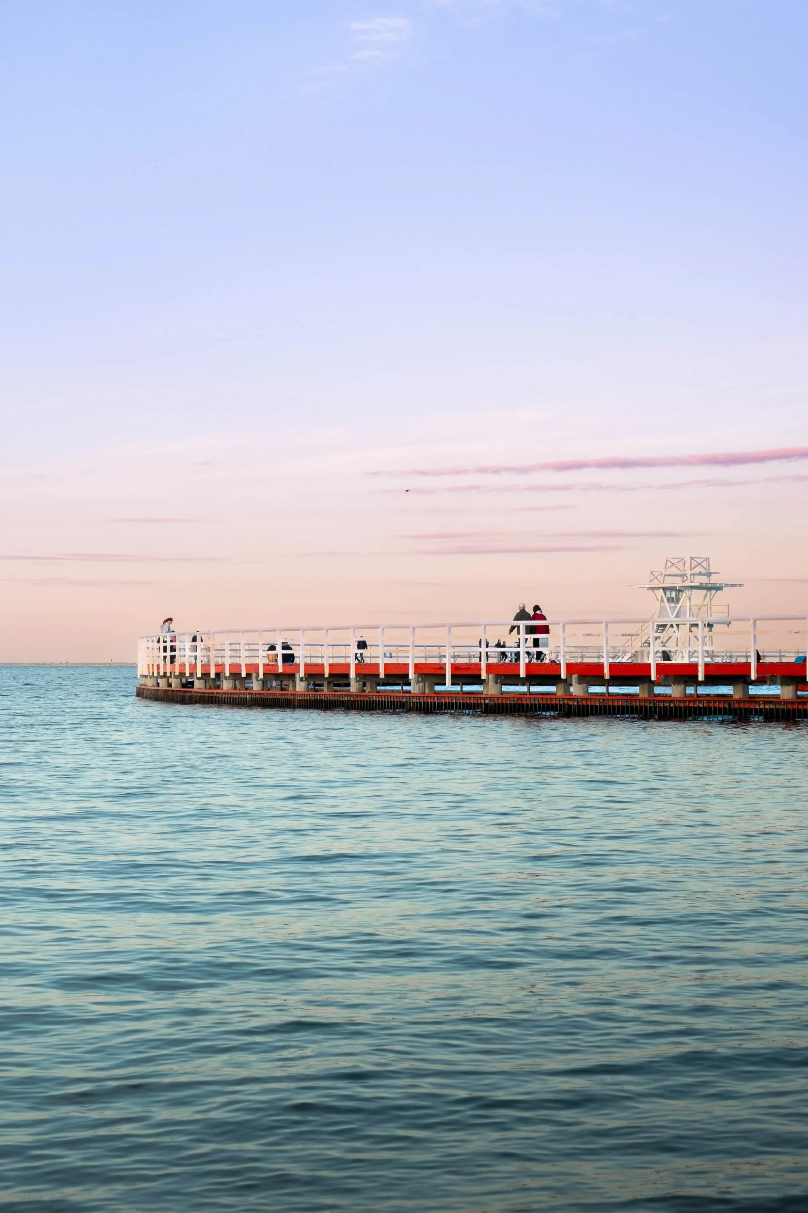 A pier extends over calm water with people walking and sitting, under a pastel-colored sky at sunset or sunrise.