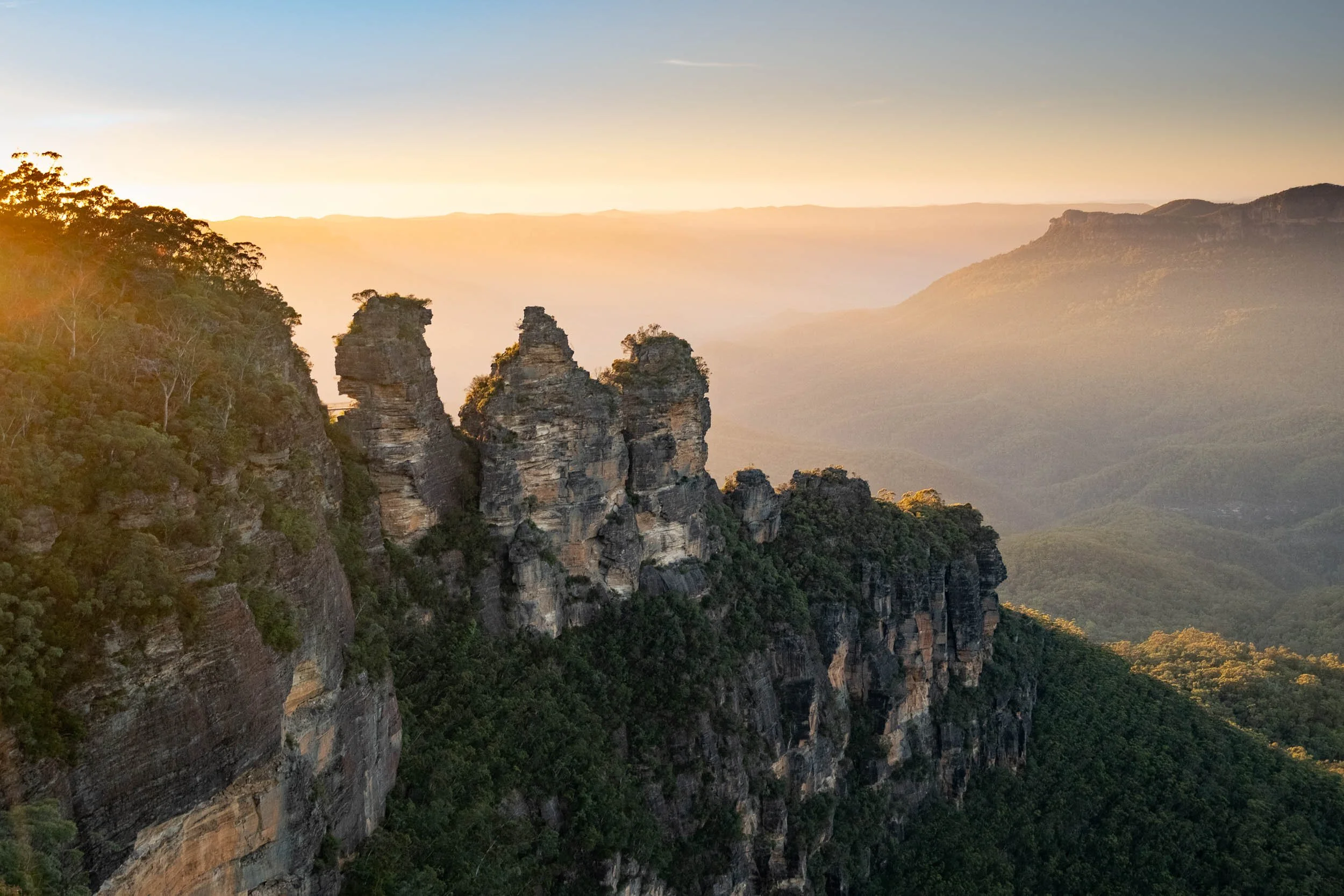 Sunrise over the Three Sisters rock formation in the Blue Mountains, Australia, with layered mountains and lush green forest canopy.